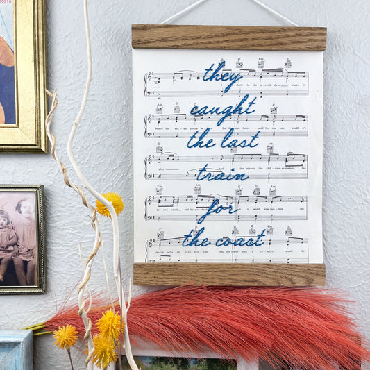sheet music for the song American Pie by Don McLean hanging in a magnetic wood frame with the words they caught the last train for the coast hand embroidered over it in blue colored thread in cursive hanging on a wall near other framed pictures art and decorative branches