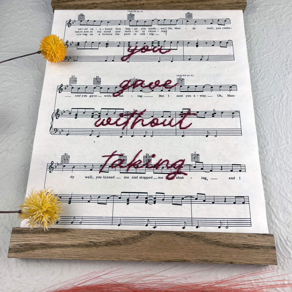 view from a low angle of sheet music for the song Mandy by Barry Manilow hanging in a magnetic wood frame with the words you gave without taking hand embroidered over it in plum colored thread in cursive hanging on a wall near botanical decorations
