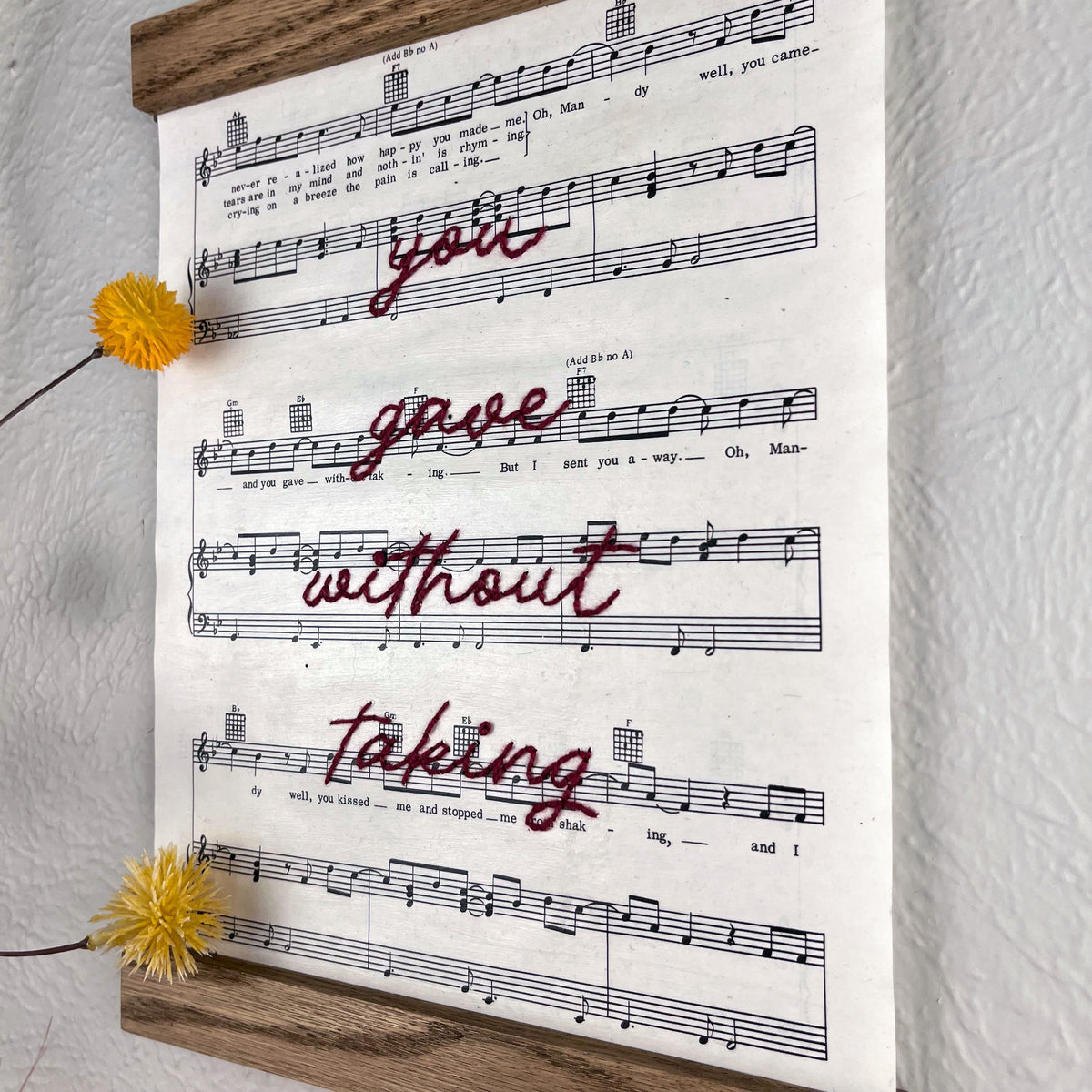 close angled view of sheet music for the song Mandy by Barry Manilow hanging in a magnetic wood frame with the words you gave without taking hand embroidered over it in plum colored thread in cursive hanging on a wall near botanical decorations