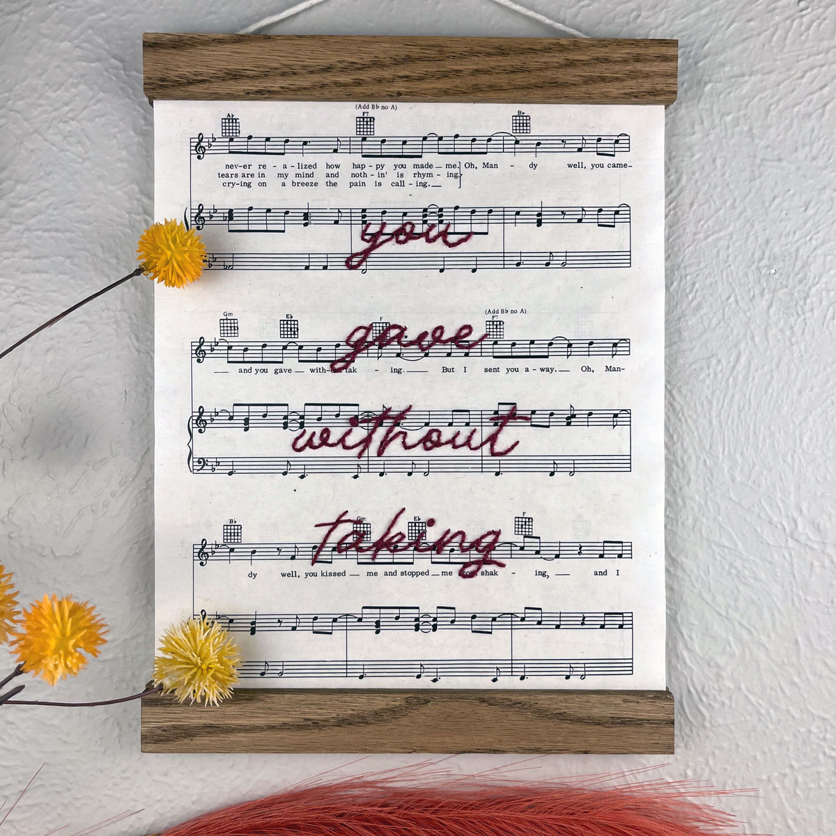 sheet music for the song Mandy by Barry Manilow hanging in a magnetic wood frame with the words you gave without taking hand embroidered over it in plum colored thread in cursive hanging on a wall near botanical decorations