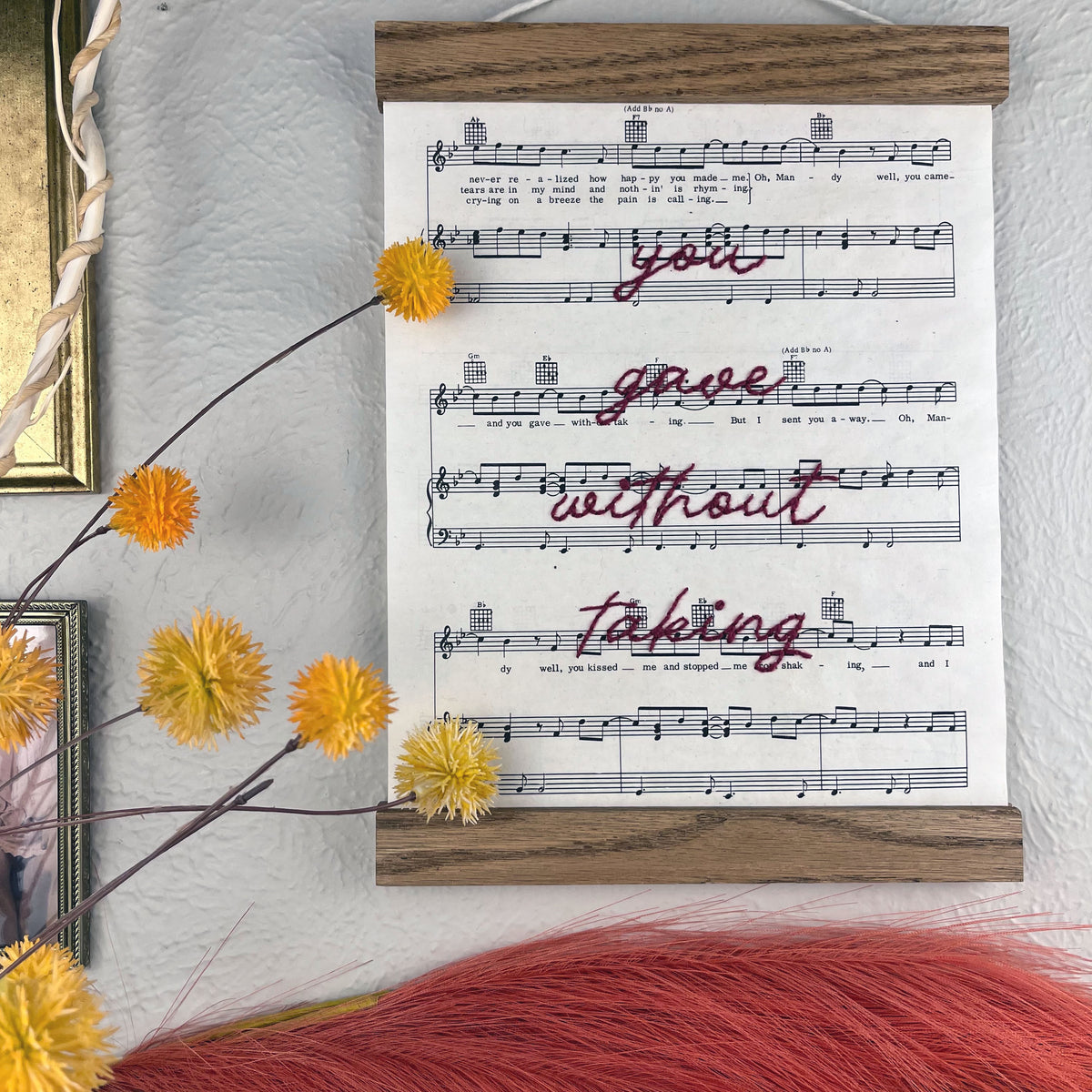sheet music for the song Mandy by Barry Manilow hanging in a magnetic wood frame with the words you gave without taking hand embroidered over it in plum colored thread in cursive hanging on a wall near botanical decorations and framed art