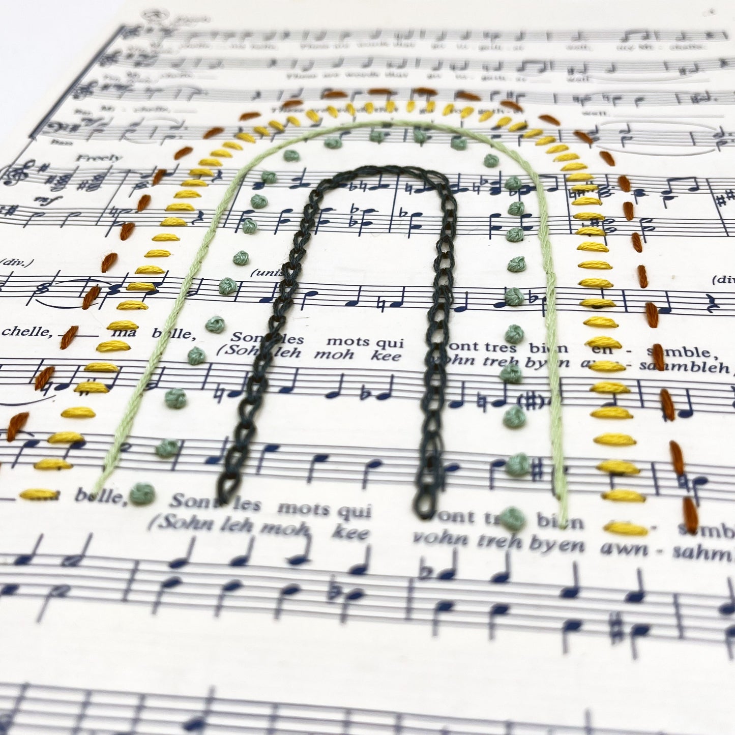 Close up angled view of sheet music for the song "Michelle", hand stitched on in the shape of a rainbow with different types of stitches like chainstitch in dark blue, french knots in green, whipped backstitch in light green, running stitch in red