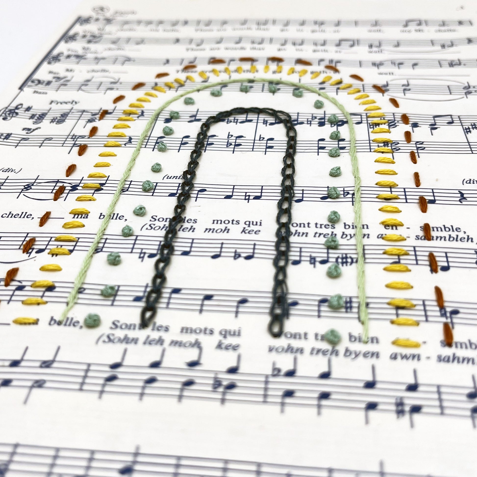 Close up angled view of sheet music for the song "Michelle", hand stitched on in the shape of a rainbow with different types of stitches like chainstitch in dark blue, french knots in green, whipped backstitch in light green, running stitch in red