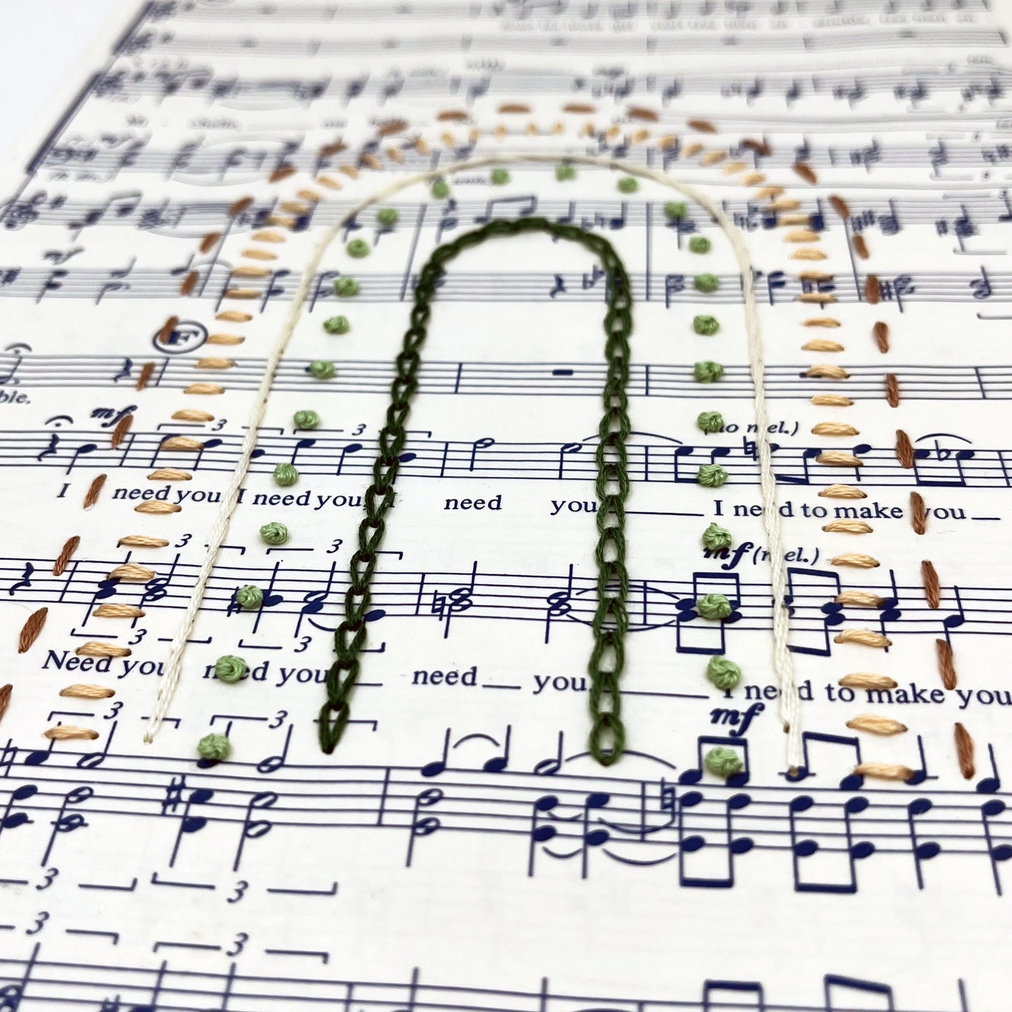 Close up angled view of sheet music for the song "Michelle", hand stitched on in the shape of a rainbow with different types of stitches like chainstitch in dark green, french knots in light green, whipped backstitch in ivory, running stitch in mauve