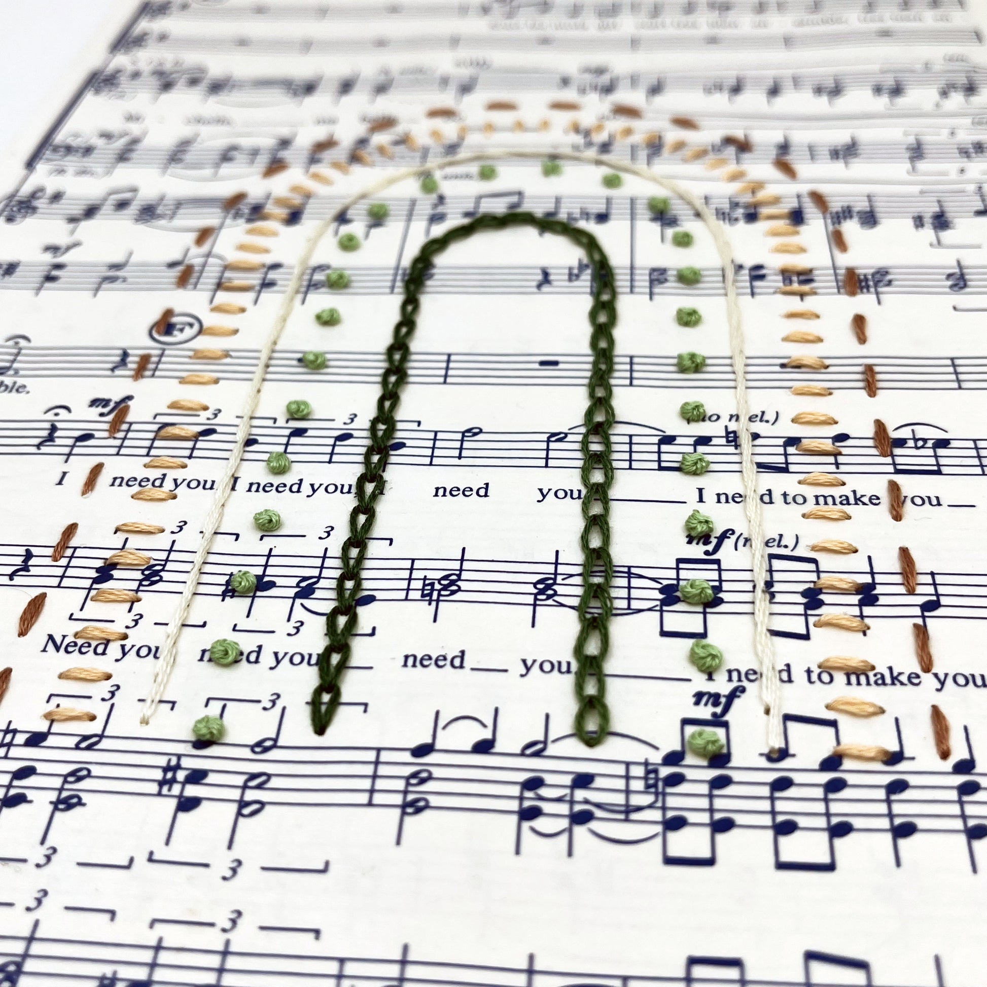 Close up angled view of sheet music for the song "Michelle", hand stitched on in the shape of a rainbow with different types of stitches like chainstitch in dark green, french knots in light green, whipped backstitch in ivory, running stitch in mauve