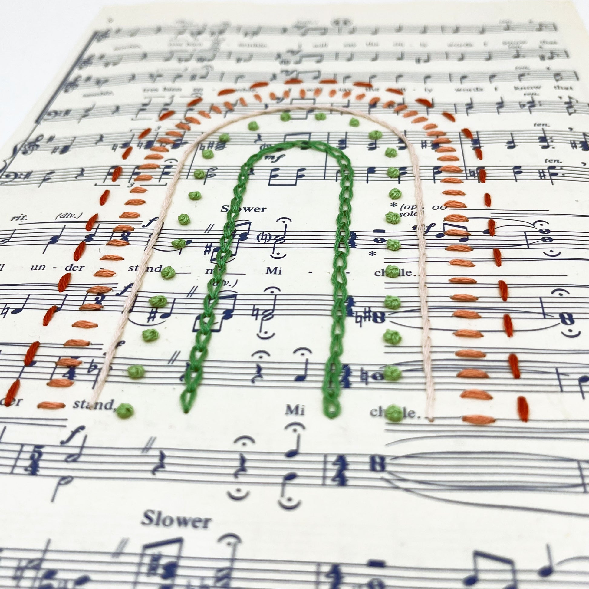 Close up angled view of sheet music for the song "Michelle", hand stitched on in the shape of a rainbow with different types of stitches like chainstitch in dark green, french knots in light green, whipped backstitch in pink, running stitch in red
