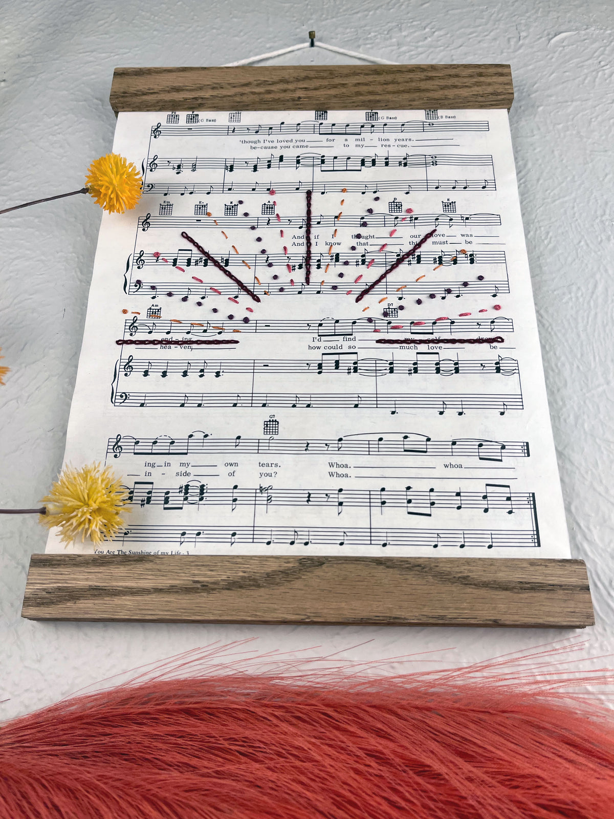 sheet music for the song You Are the Sunshine of my Life by Stevie Wonder hanging in a magnetic wood frame with rays from a setting sun hand embroidered over it in a variety of stitches and warm colors hanging on a wall near botanical decorations