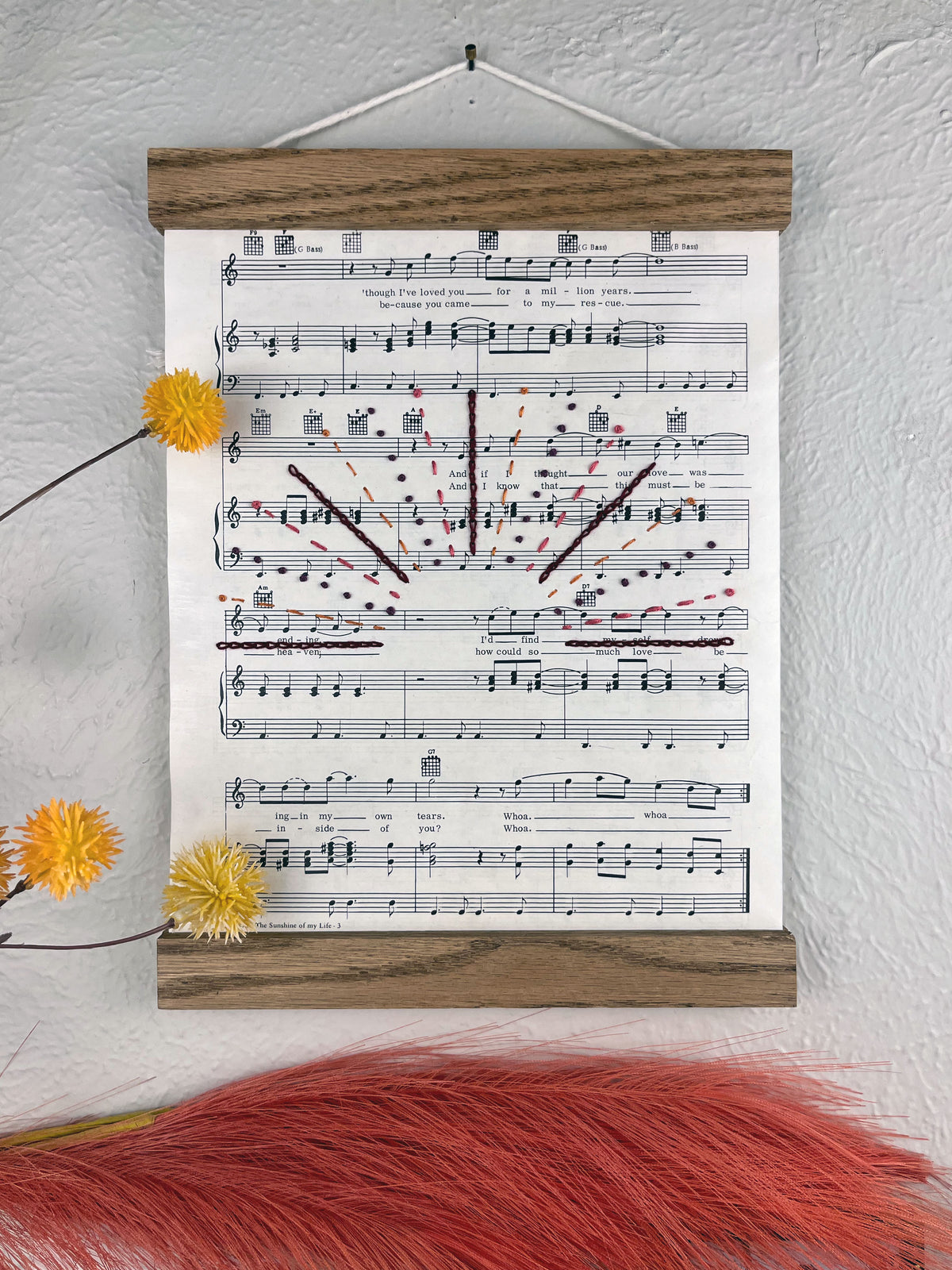sheet music for the song You Are the Sunshine of my Life by Stevie Wonder hanging in a magnetic wood frame with rays from a setting sun hand embroidered over it in a variety of stitches and warm colors hanging on a wall near botanical decorations