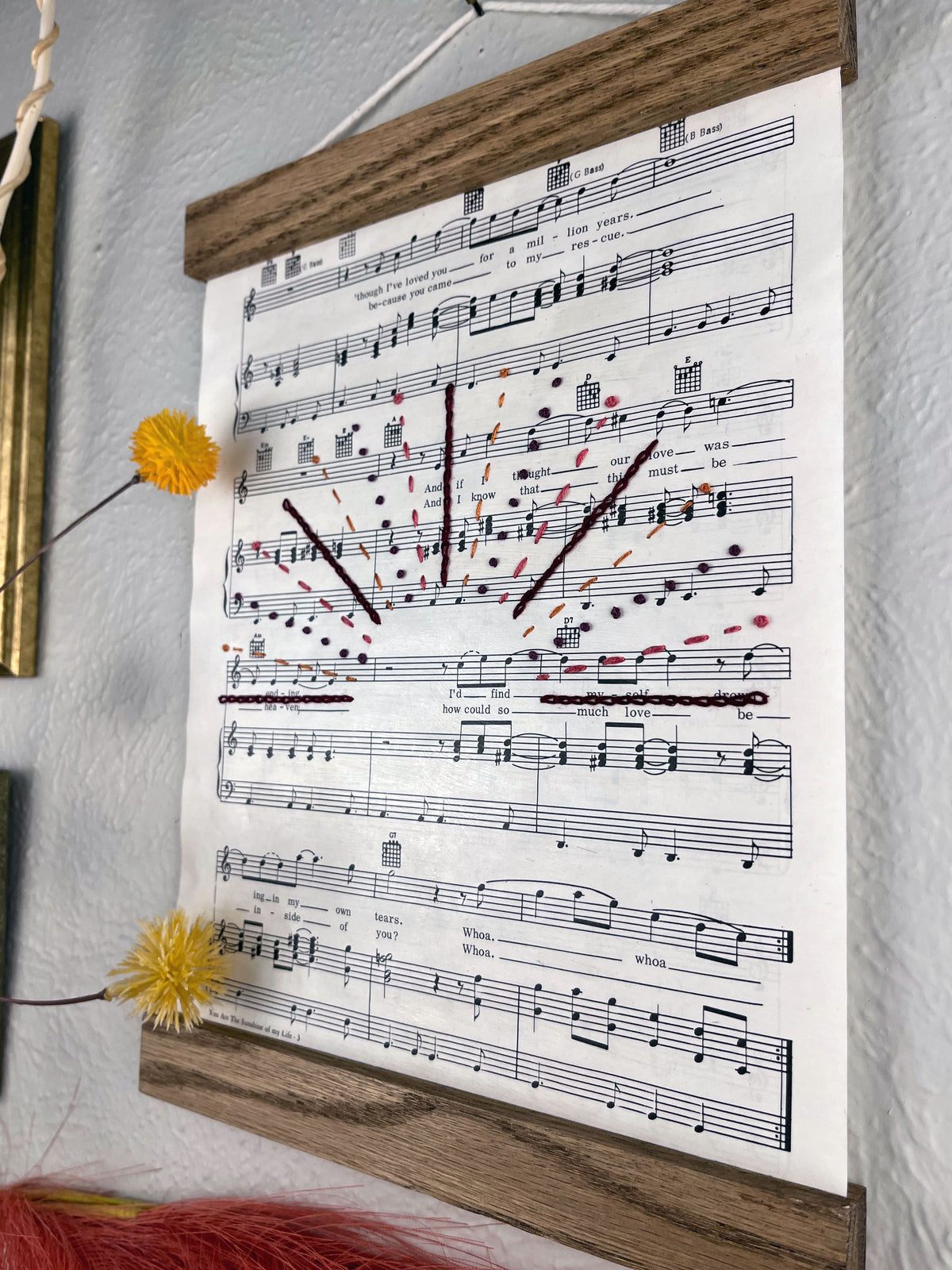 angled view of sheet music for the song You Are the Sunshine of my Life by Stevie Wonder hanging in a magnetic wood frame with rays from a setting sun hand embroidered over it in a variety of stitches and warm colors hanging on a wall near botanical decorations and framed art