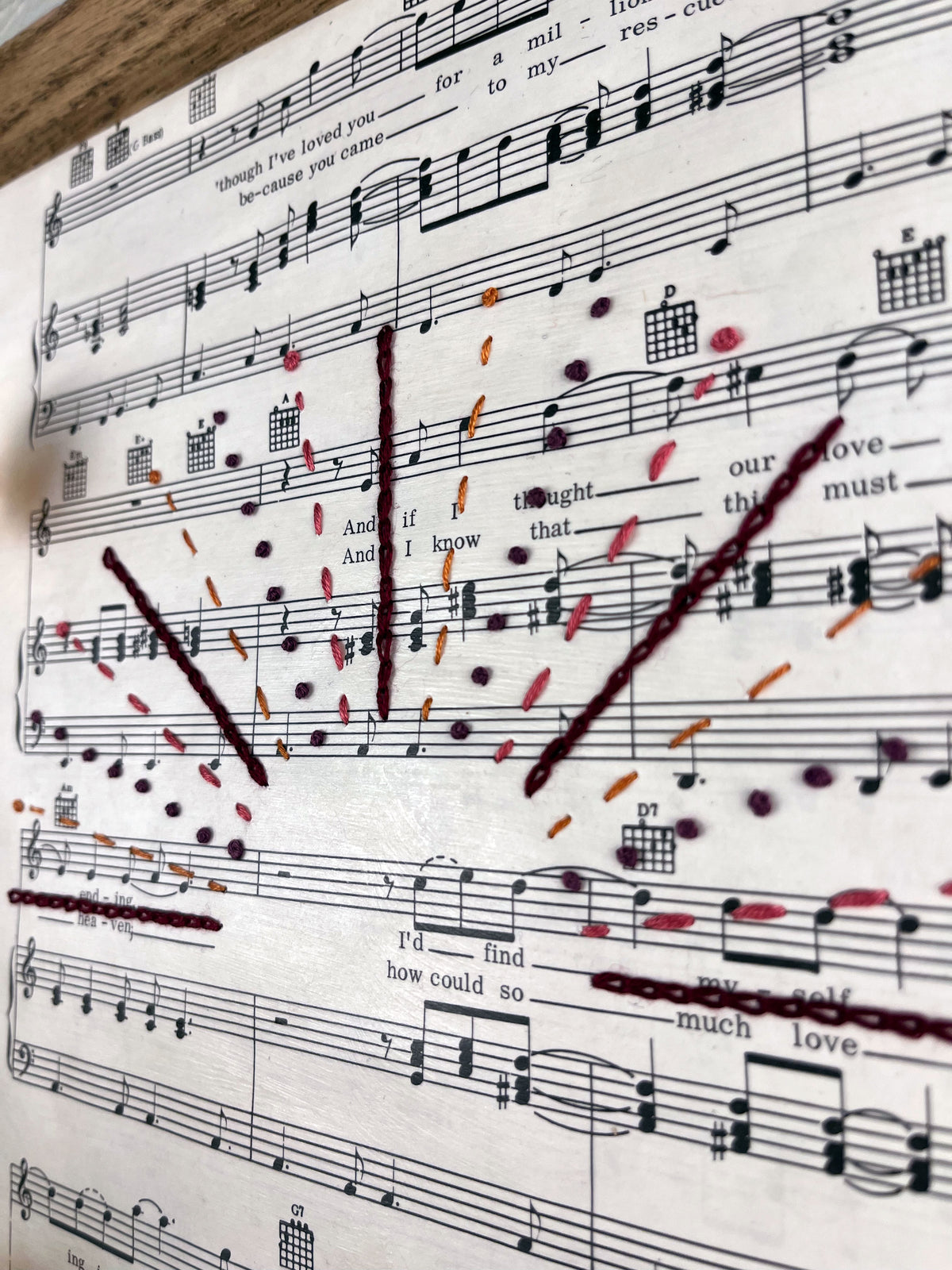 close up view of sheet music for the song You Are the Sunshine of my Life by Stevie Wonder hanging in a magnetic wood frame with rays from a setting sun hand embroidered over it in a variety of stitches and warm colors 