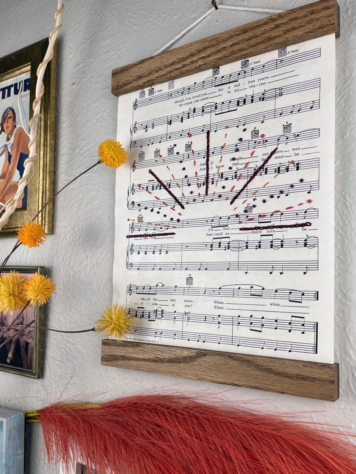 angled view of sheet music for the song You Are the Sunshine of my Life by Stevie Wonder hanging in a magnetic wood frame with rays from a setting sun hand embroidered over it in a variety of stitches and warm colors hanging on a wall near botanical decorations and framed art