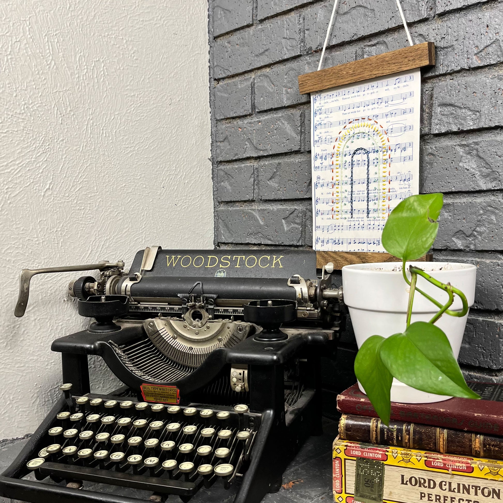 Sheet music for the song "Michelle", hand stitched on with a rainbow with different types of stitches like chainstitch, running stitch and french knots, hanging in a wood magnetic frame on a grey brick wall, with a plant and typewriter in front of it