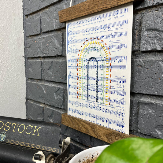 Sheet music for the song "Michelle", hand stitched on with a rainbow with different types of stitches like chainstitch, running stitch and french knots, hanging in a wood magnetic frame on a grey brick wall, with a plant and typewriter in front of it