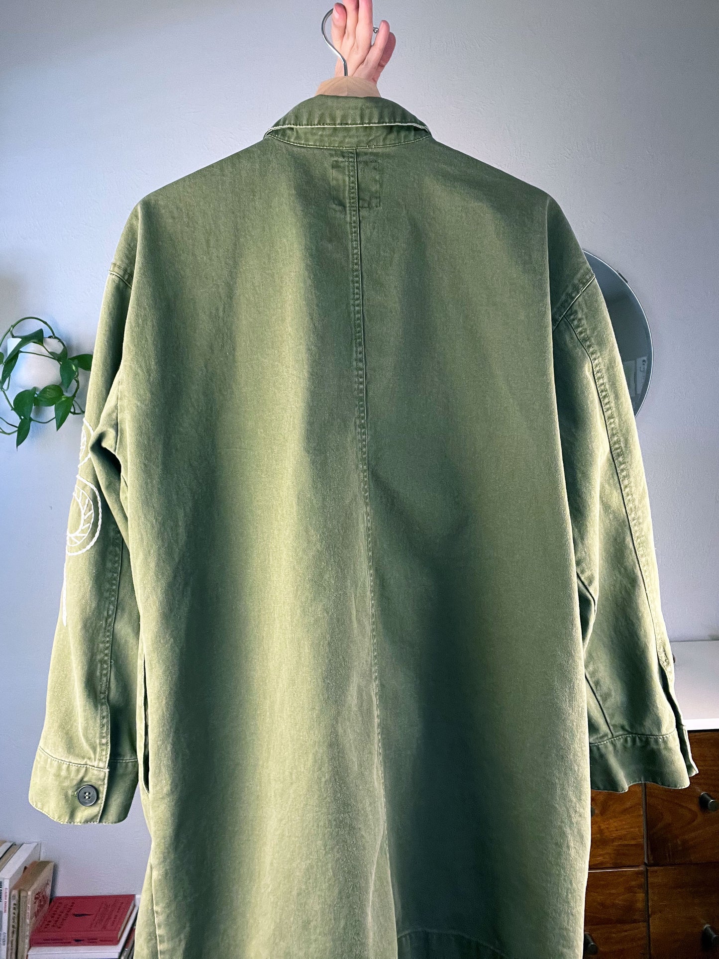 back view of an olive green jacket being held up on a hanger with a dresser plant and books in the background