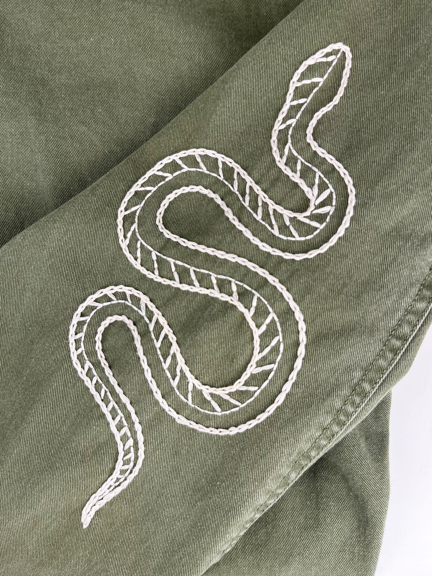 close view of a hand embroidered snake on the sleeve of a long oversized olive green jacket on a white counter