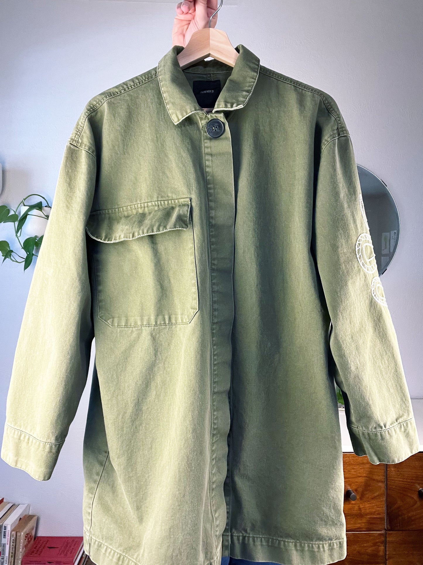 front view of an olive green jacket being held up on a hanger with a dresser plant and books in the background