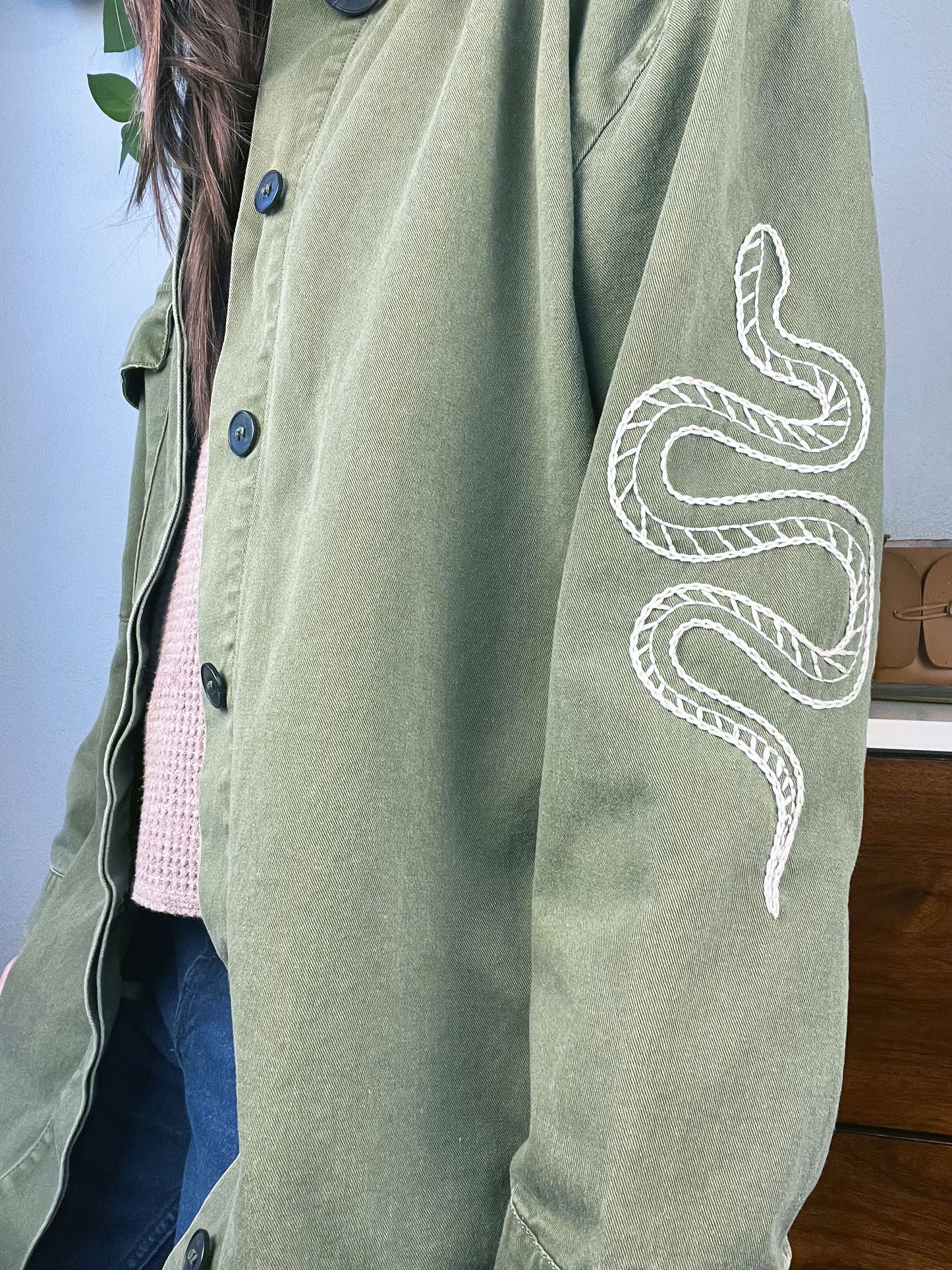 close up angled side view of a woman wearing a long oversized olive green jacket with a snake hand embroidered on the sleeve