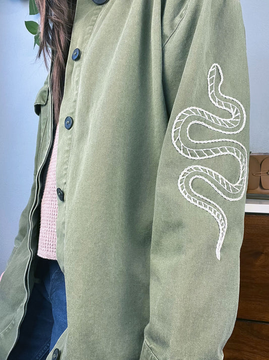 close up angled side view of a woman wearing a long oversized olive green jacket with a snake hand embroidered on the sleeve