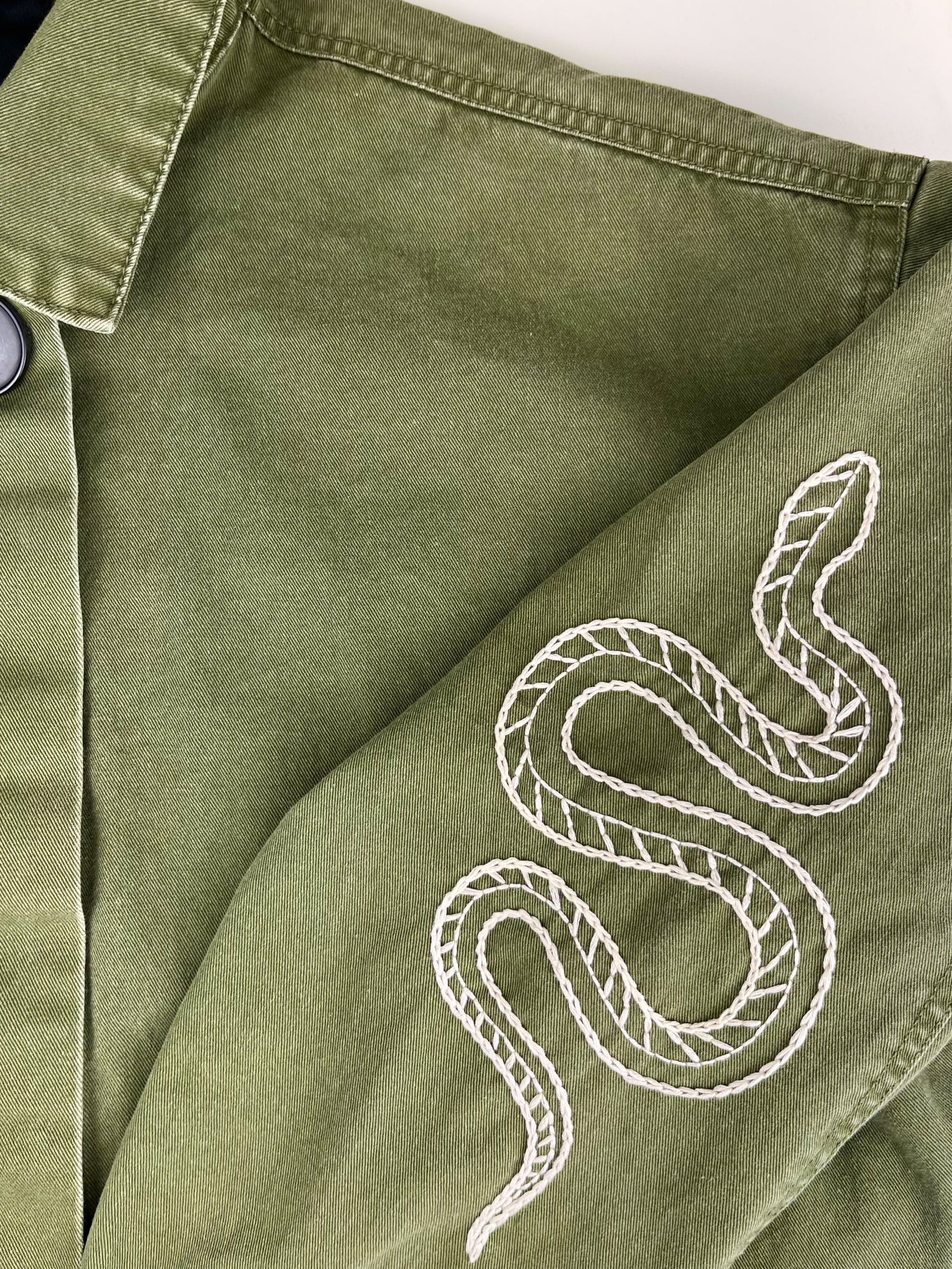 close view of a hand embroidered snake on the sleeve of a long oversized olive green jacket on a white counter