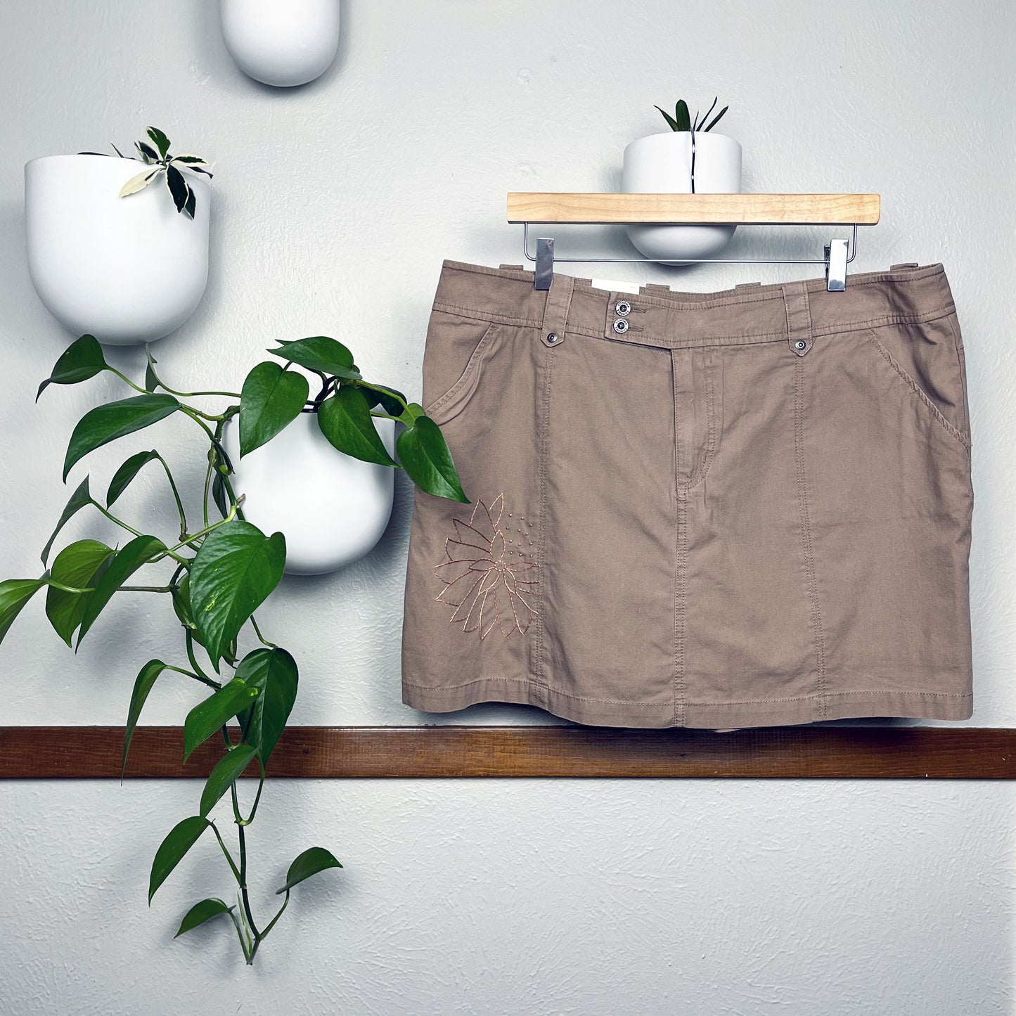 a mauve skirt on a hanger, the left side of the skirt has a hand embroidered dandelion in the same color as the skirt, hanging on a wall next to a pothos plant in a white round planter