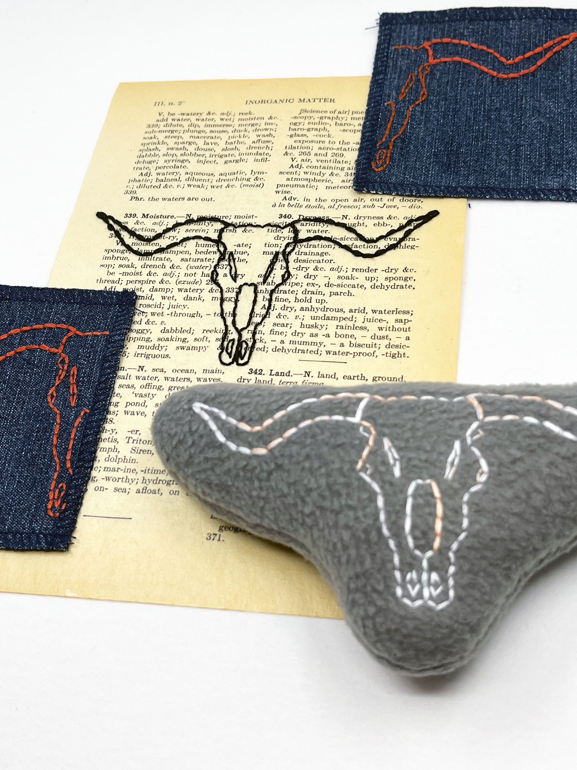 a group of things including a vintage thesaurus page, two denim patches and a grey stuffie hand embroidered with a longhorn skull