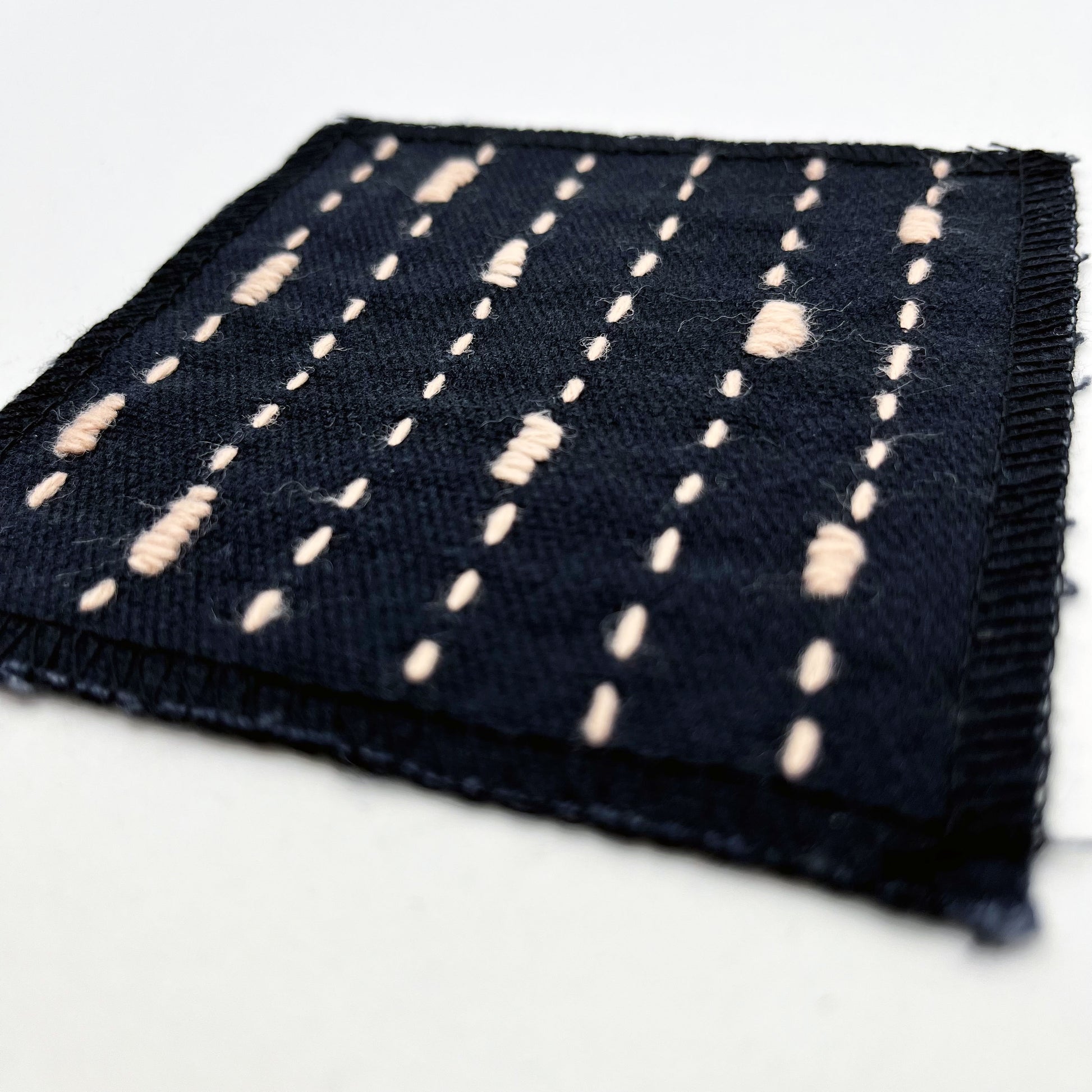 a close up angled view of a square patch made out of black canvas, handstitched with peach running stitches with randomly placed small rectangles made of stitches running the other direction, with overlocked edges, on a white background