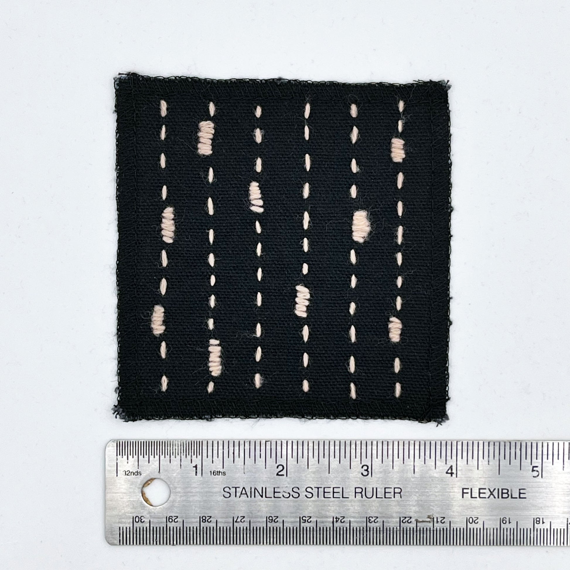 a square patch made out of black canvas, handstitched with peach running stitches with randomly placed small rectangles made of stitches running the other direction, with overlocked edges, on a white background next to a ruler to show width of 4 in