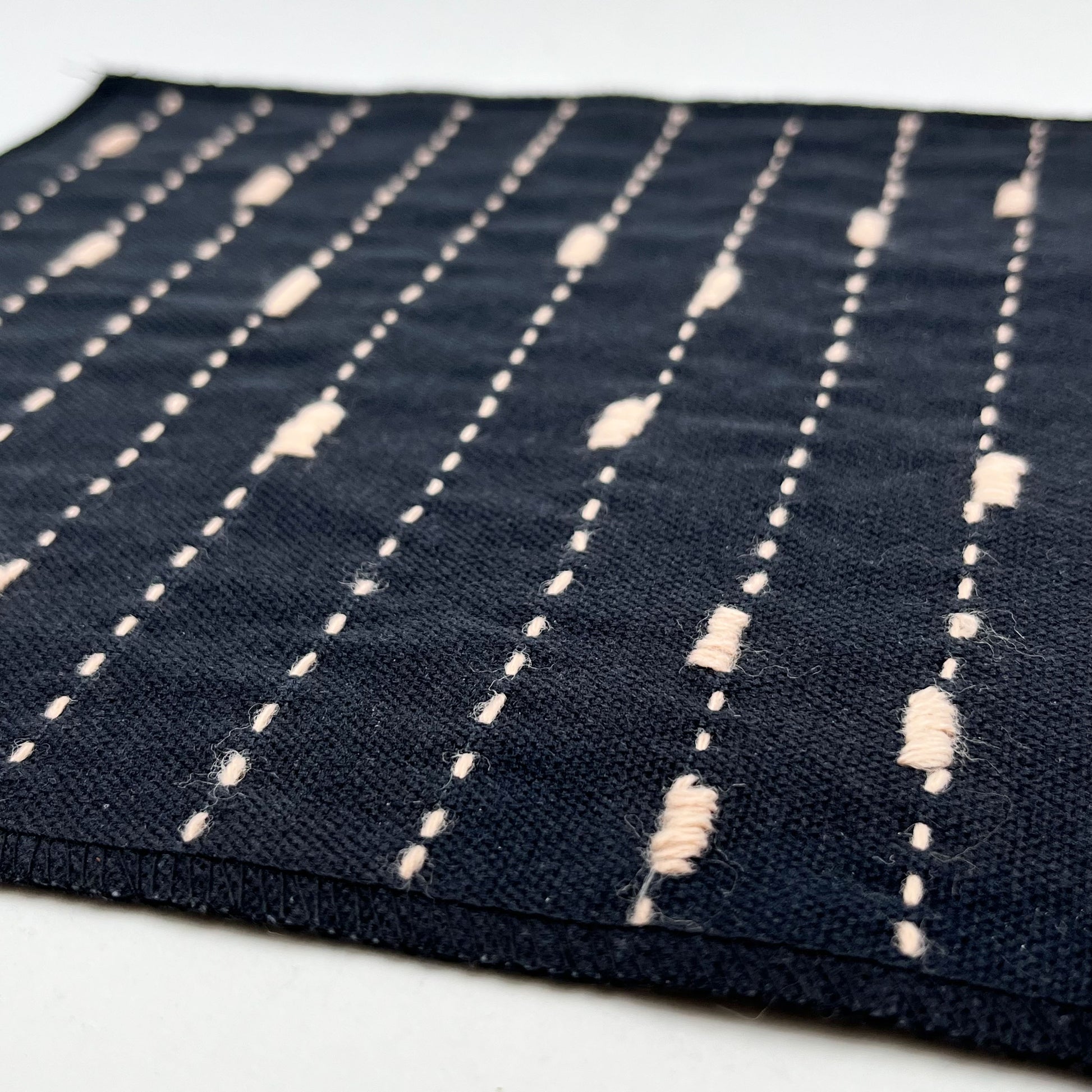 a close up angled view of a square patch made out of black canvas, handstitched with peach running stitches with randomly placed small rectangles made of stitches running the other direction, with overlocked edges, on a white background