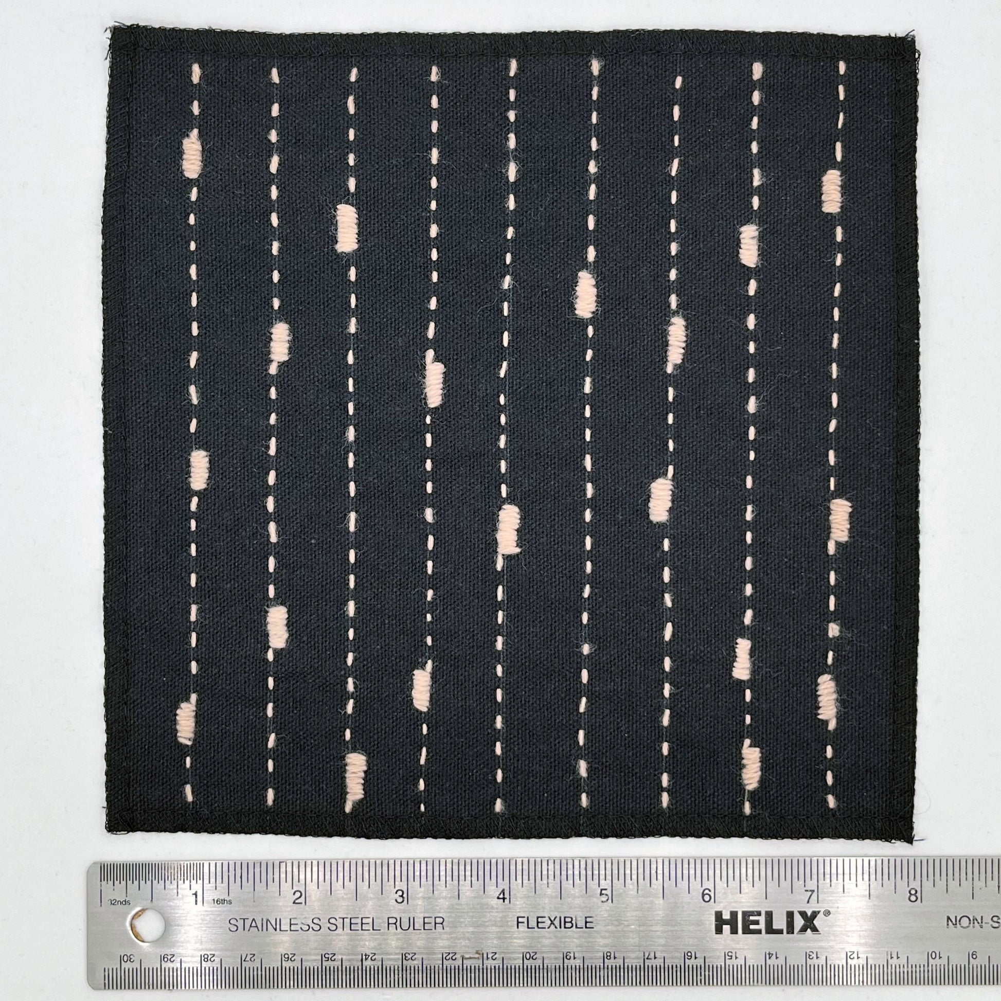 a square patch made out of black canvas, handstitched with peach running stitches with randomly placed small rectangles made of stitches running the other direction, with overlocked edges, on a white background next to a ruler to show width of 8 in