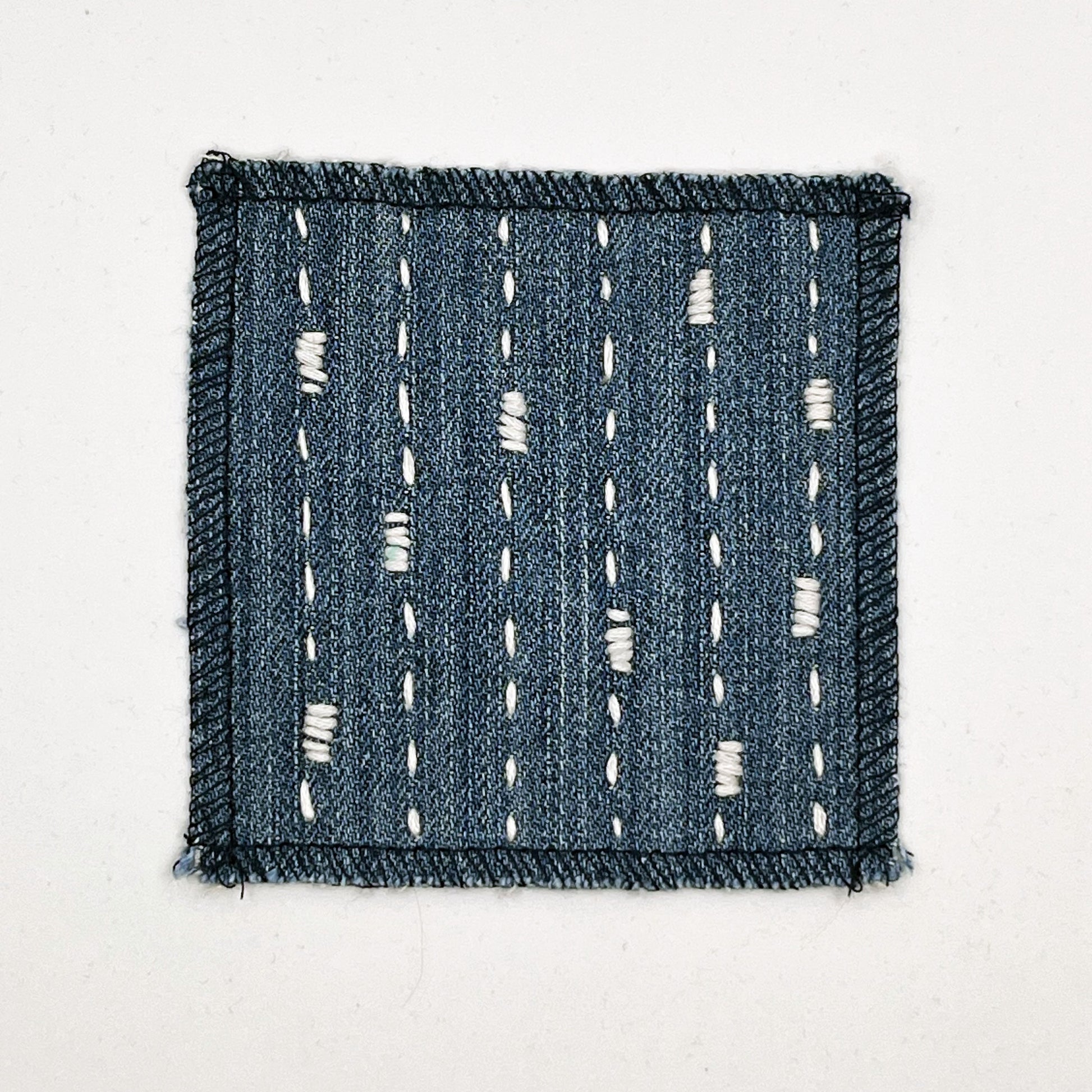 a square patch made out of denim, handstitched with ivory running stitches with randomly placed small rectangles made of stitches running the other direction, with overlocked edges, on a white background