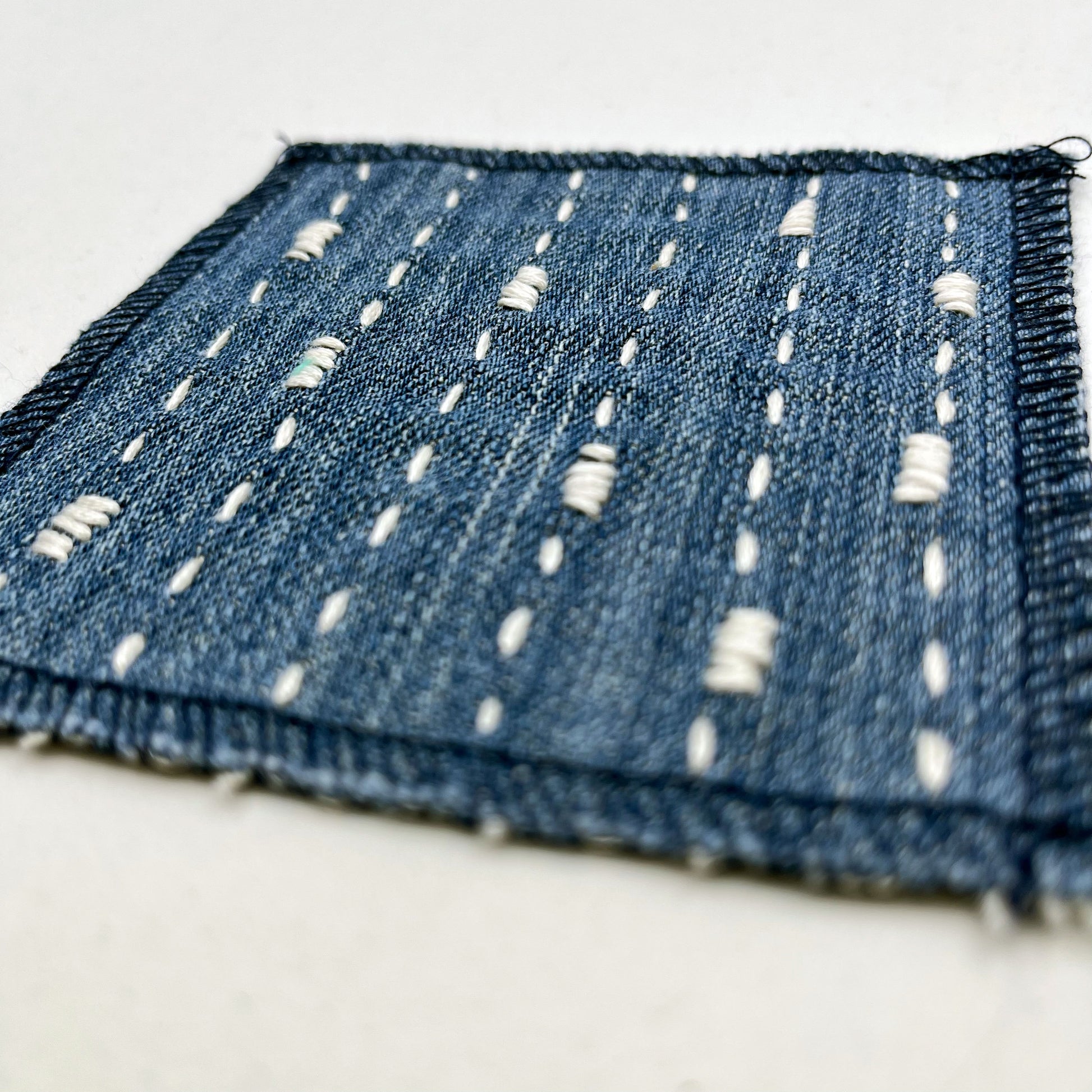 close up angled view of a square patch made out of denim, handstitched with ivory running stitches with randomly placed small rectangles made of stitches running the other direction, with overlocked edges, on a white background