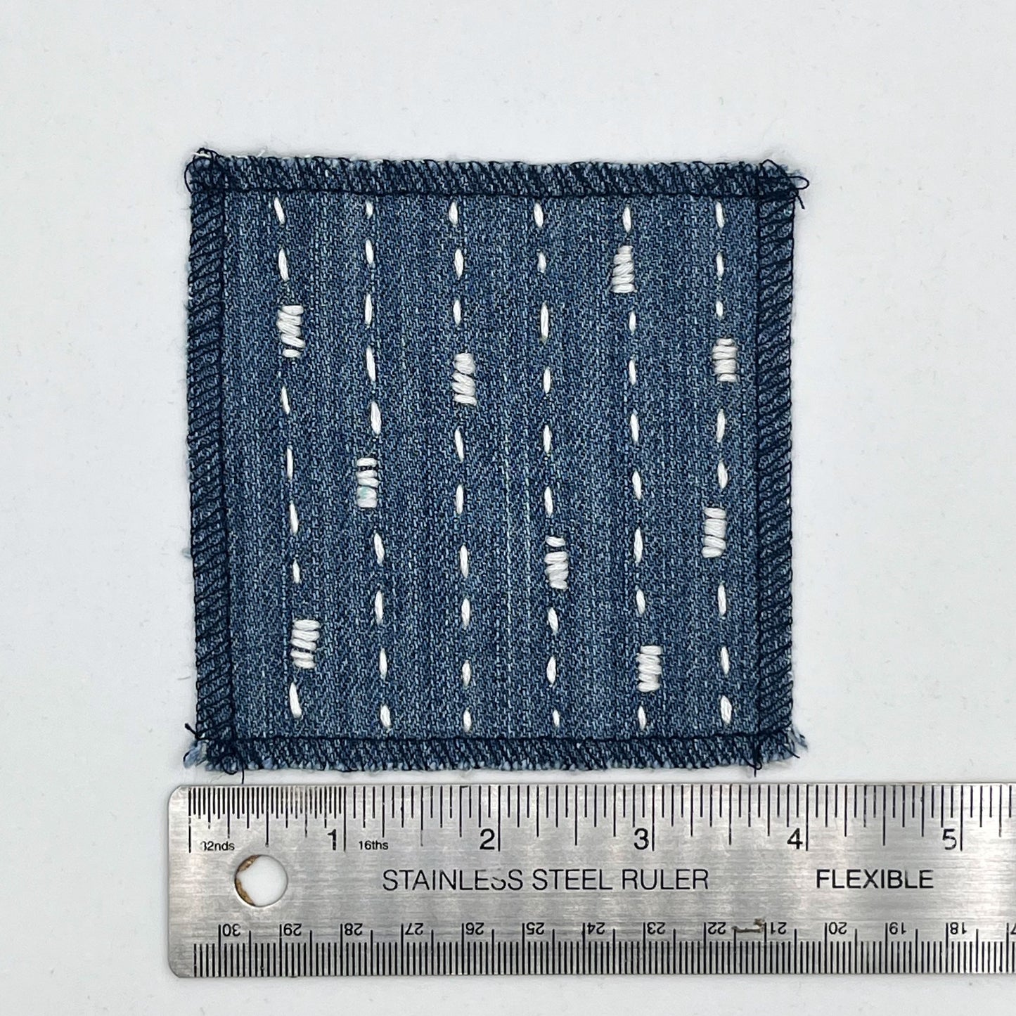 a square patch made out of denim, handstitched with ivory running stitches with randomly placed small rectangles made of stitches running the other direction, with overlocked edges, on a white background next to a ruler to show width of 4 inches