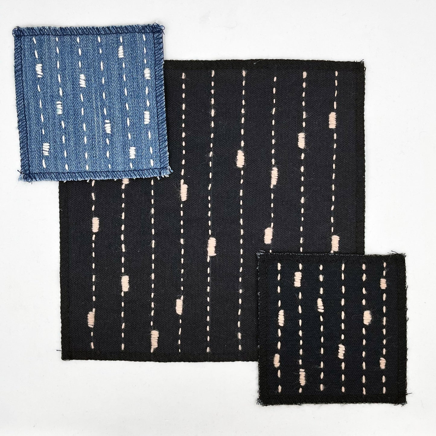 a group of 3 square patches made out of denim and black canvas, handstitched in ivory (on the denim) and peach (on the black) running stitches with randomly placed small rectangles made of stitches running the other direction, on a white background
