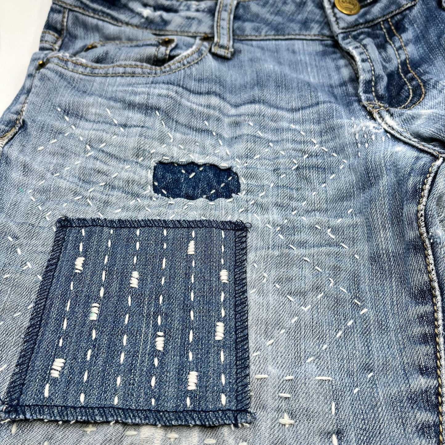 a square patch made out of denim, handstitched with ivory running stitches with randomly placed small rectangles made of stitches running the other direction, on a pair of jeans with other visible mends