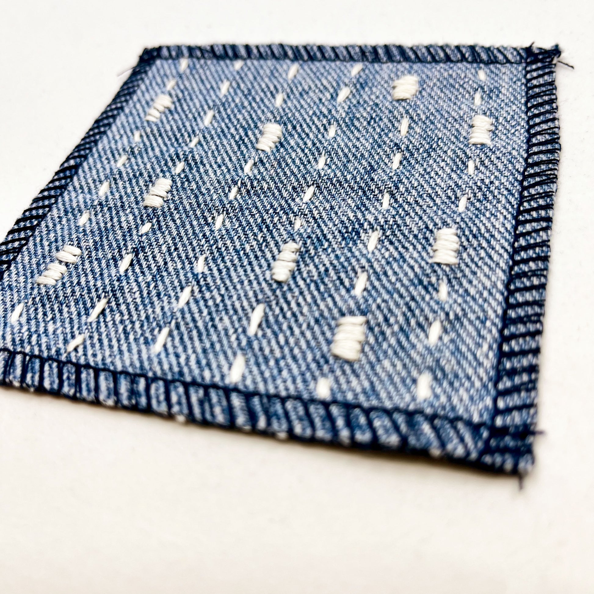 close up angled view of a square patch made out of denim, handstitched with ivory running stitches with randomly placed small rectangles made of stitches running the other direction, with overlocked edges, on a white background