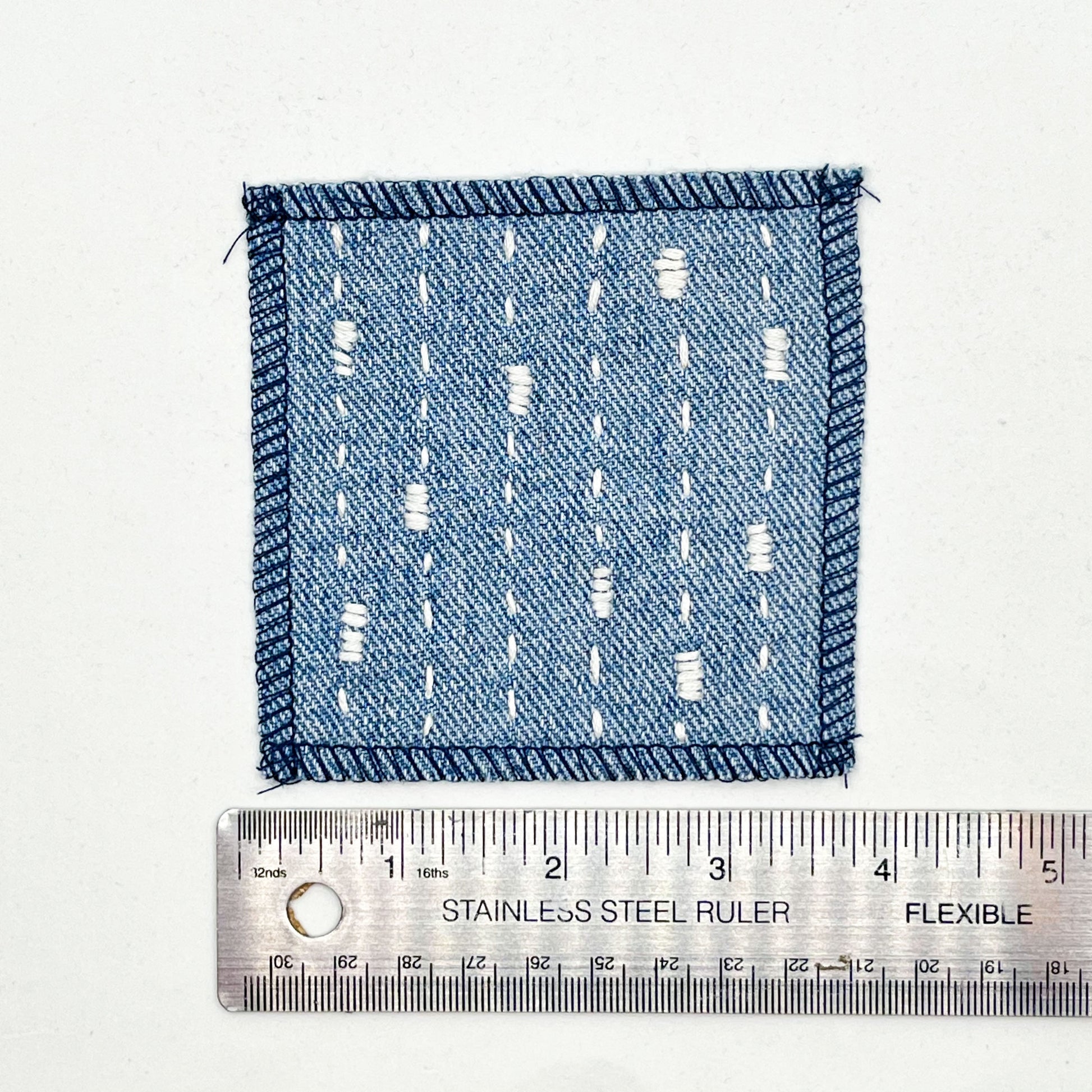 a square patch made out of denim, handstitched with ivory running stitches with randomly placed small rectangles made of stitches running the other direction, with overlocked edges, on a white background, placed next to a metal ruler to show a width of four inches