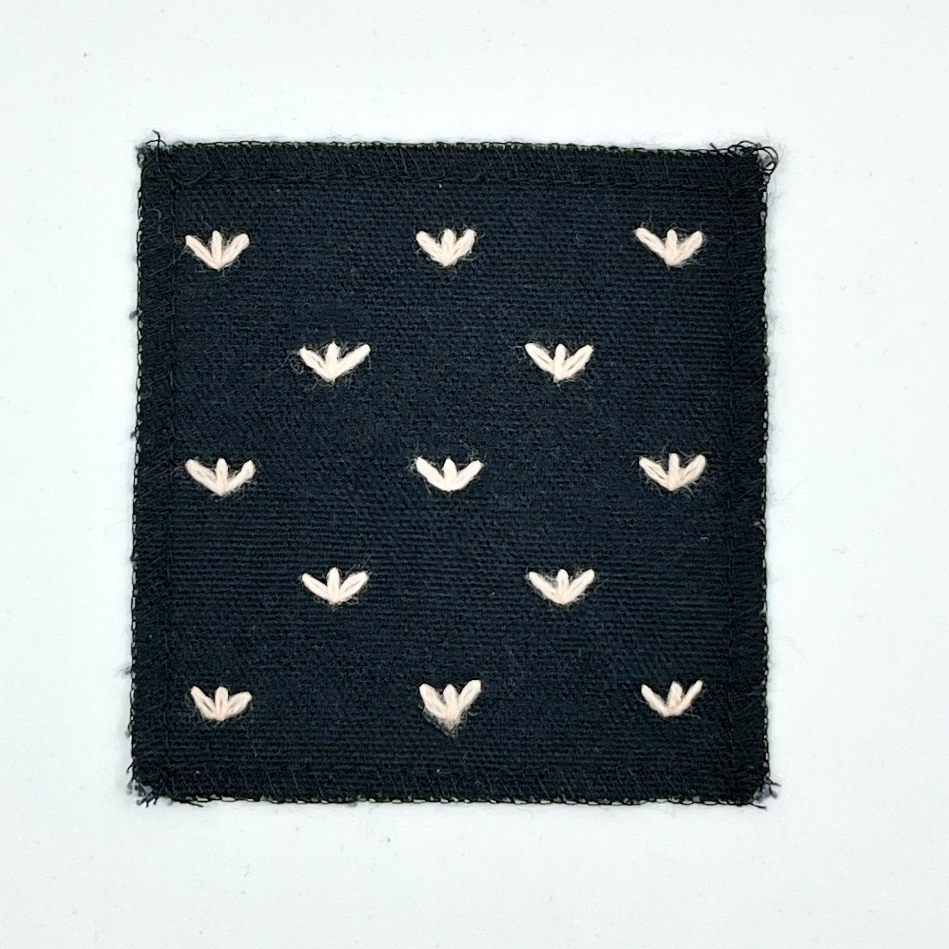 a square black canvas patch, with peach stitches that look like birds feet or sprouts spread out in a diamond pattern, with overlocked edges, on a white background