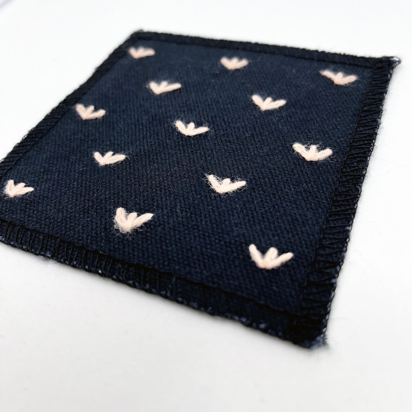 a close up angled view of a square black canvas patch, with peach stitches that look like birds feet or sprouts spread out in a diamond pattern, with overlocked edges, on a white background