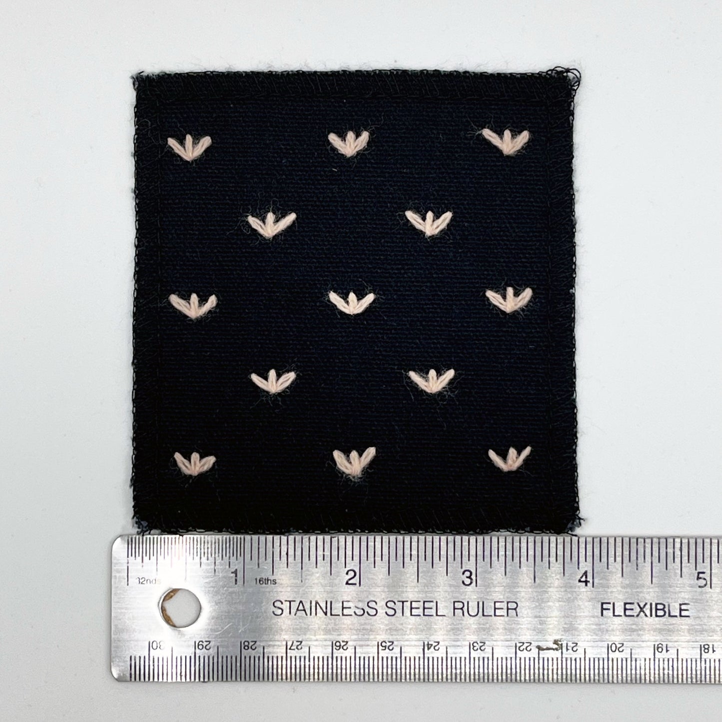a square black canvas patch, with peach stitches that look like birds feet or sprouts spread out in a diamond pattern, with overlocked edges, placed next to a metal ruler to show a width of four inches, on a white background