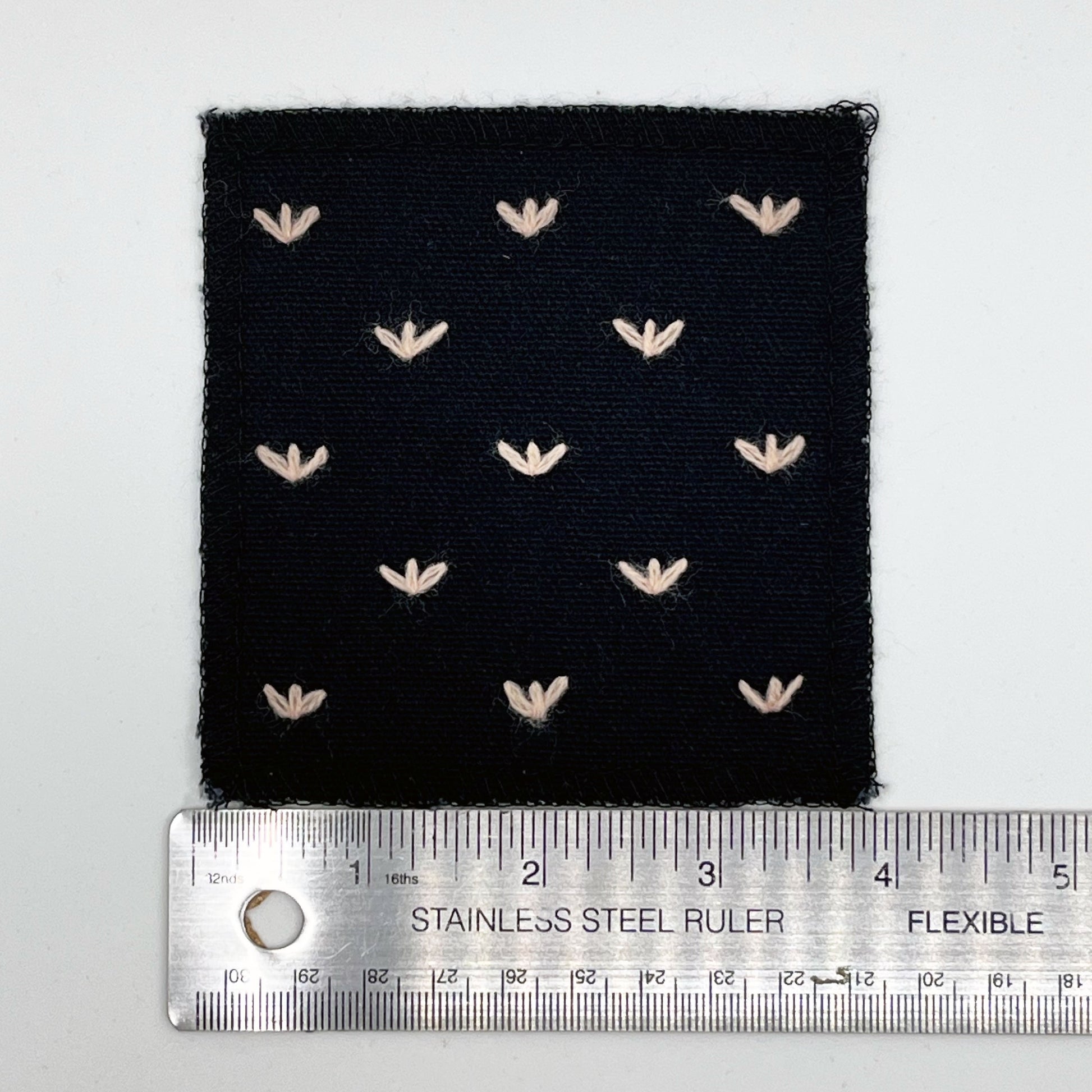 a square black canvas patch, with peach stitches that look like birds feet or sprouts spread out in a diamond pattern, with overlocked edges, placed next to a metal ruler to show a width of four inches, on a white background