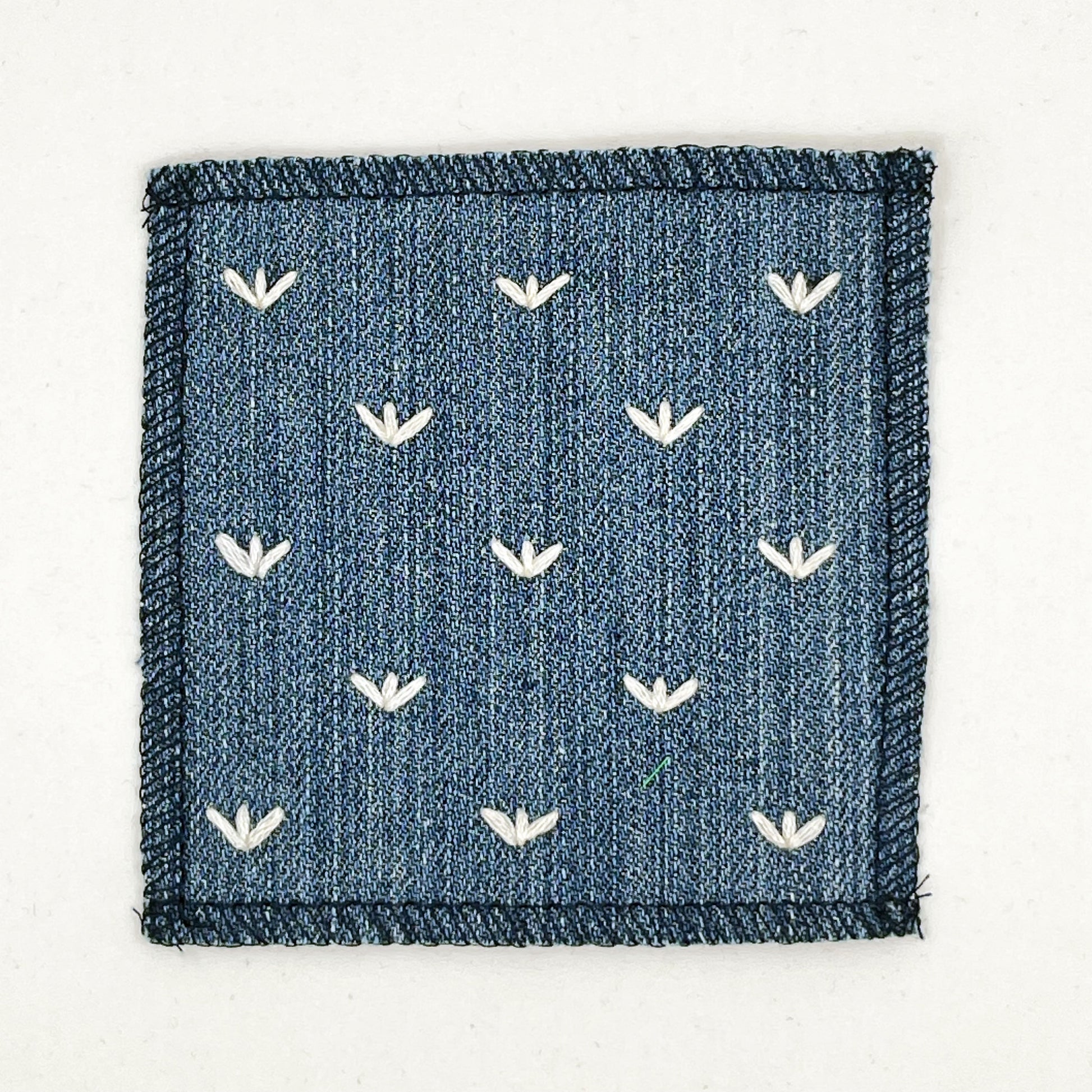 a square denim patch, with ivory stitches that look like birds feet or sprouts spread out in a diamond pattern, with overlocked edges, on a white background