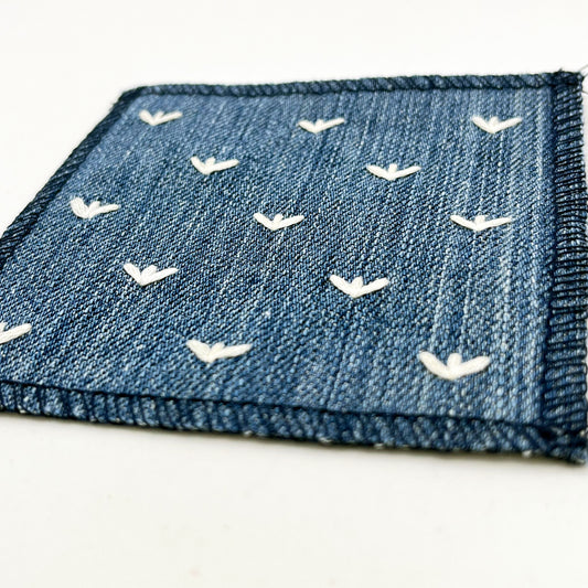 a close up angled view of a square denim patch, with ivory stitches that look like birds feet or sprouts spread out in a diamond pattern, with overlocked edges, on a white background