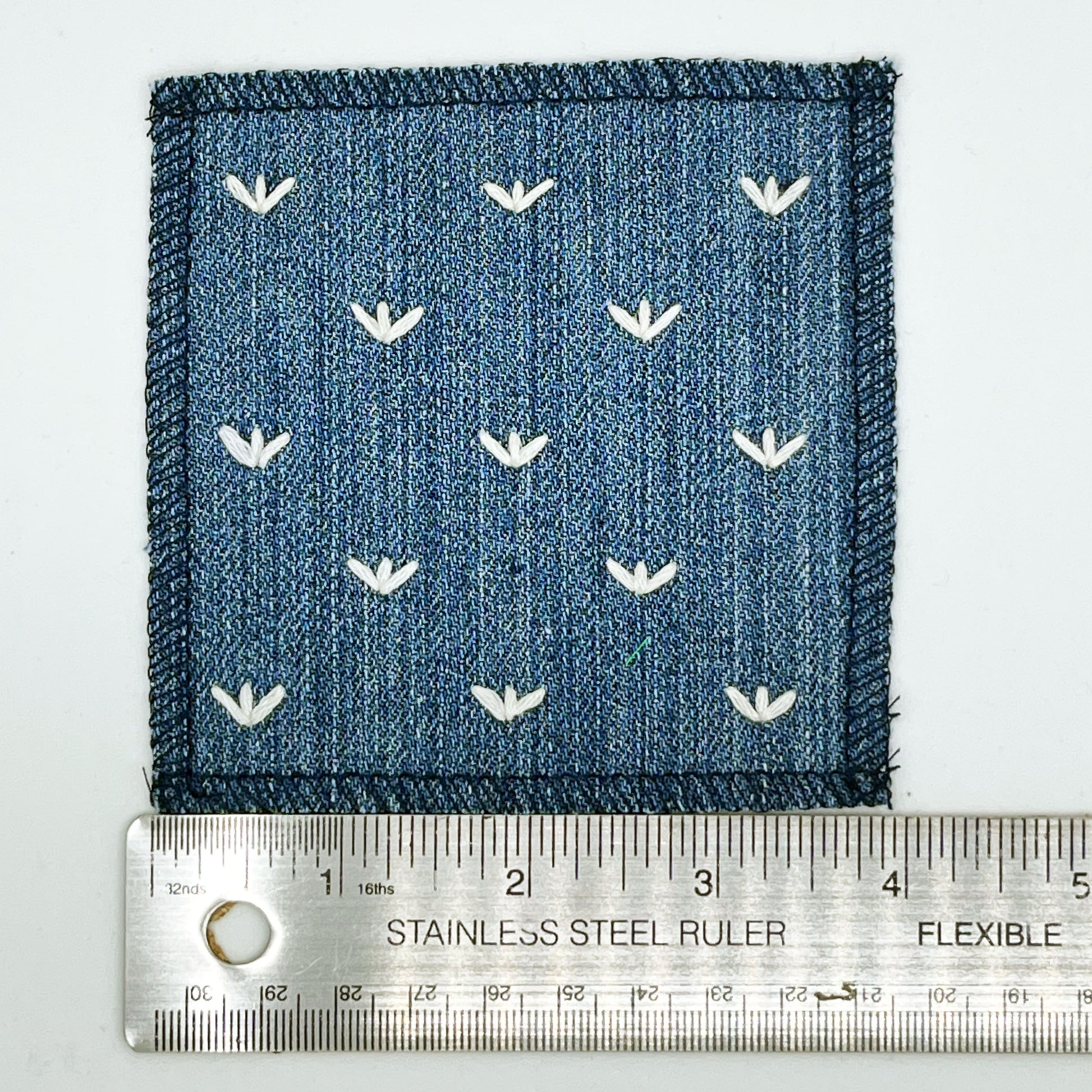 a square denim patch, with ivory stitches that look like birds feet or sprouts spread out in a diamond pattern, with overlocked edges, placed next to a metal ruler to show a width of four inches