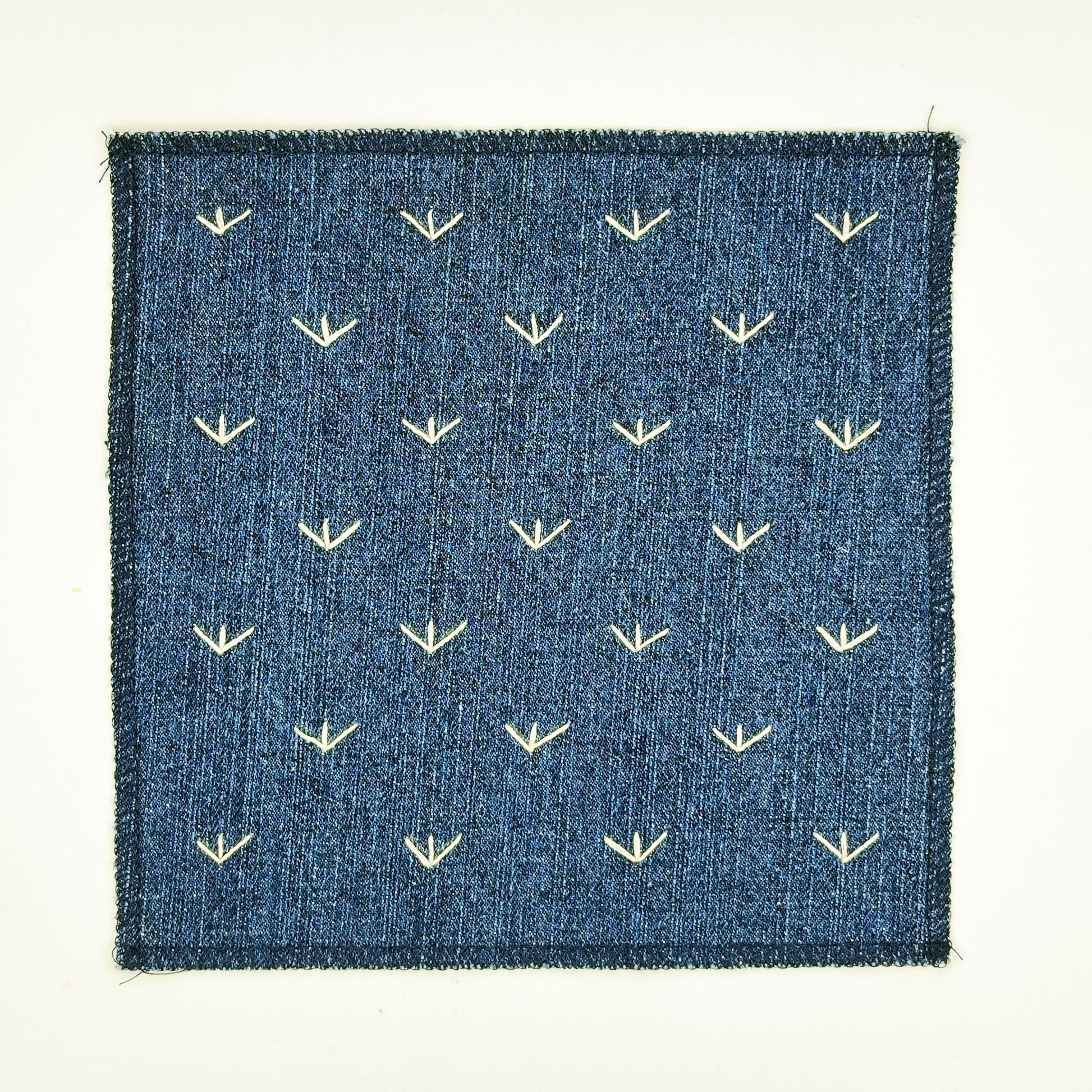 a square denim patch, with ivory stitches that look like birds feet or sprouts spread out in a diamond pattern, with overlocked edges, on a white background