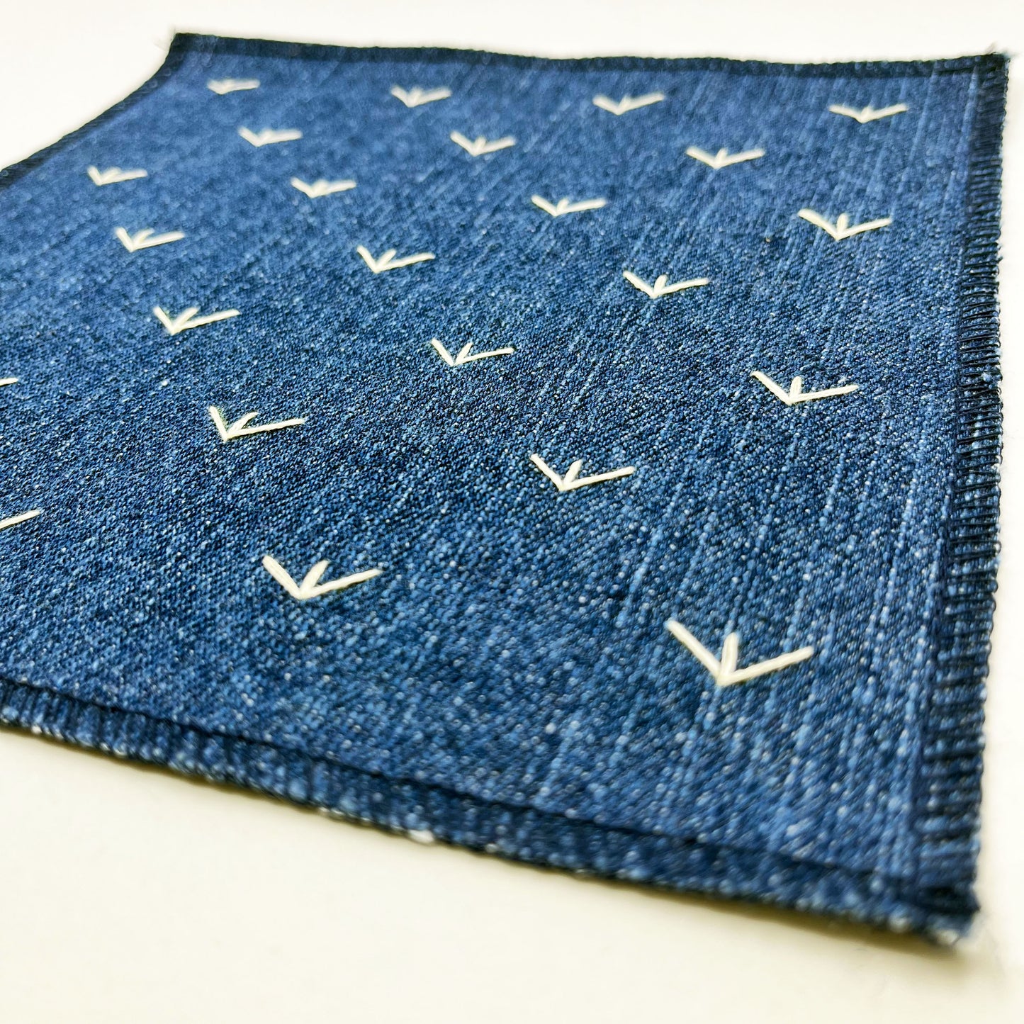 a close up angled view of a square denim patch, with ivory stitches that look like birds feet or sprouts spread out in a diamond pattern, with overlocked edges, on a white background