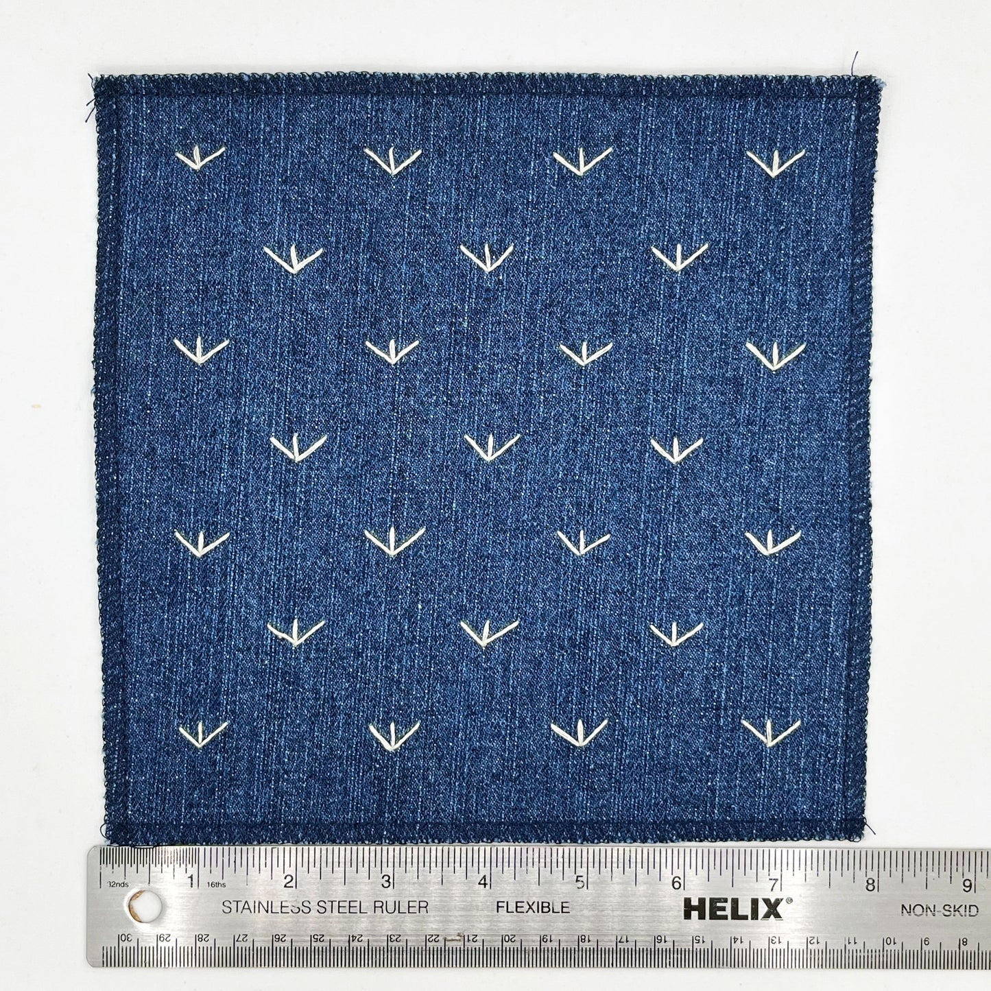 a square denim patch, with ivory stitches that look like birds feet or sprouts spread out in a diamond pattern, with overlocked edges, placed next to a metal ruler to show a width of eight inches