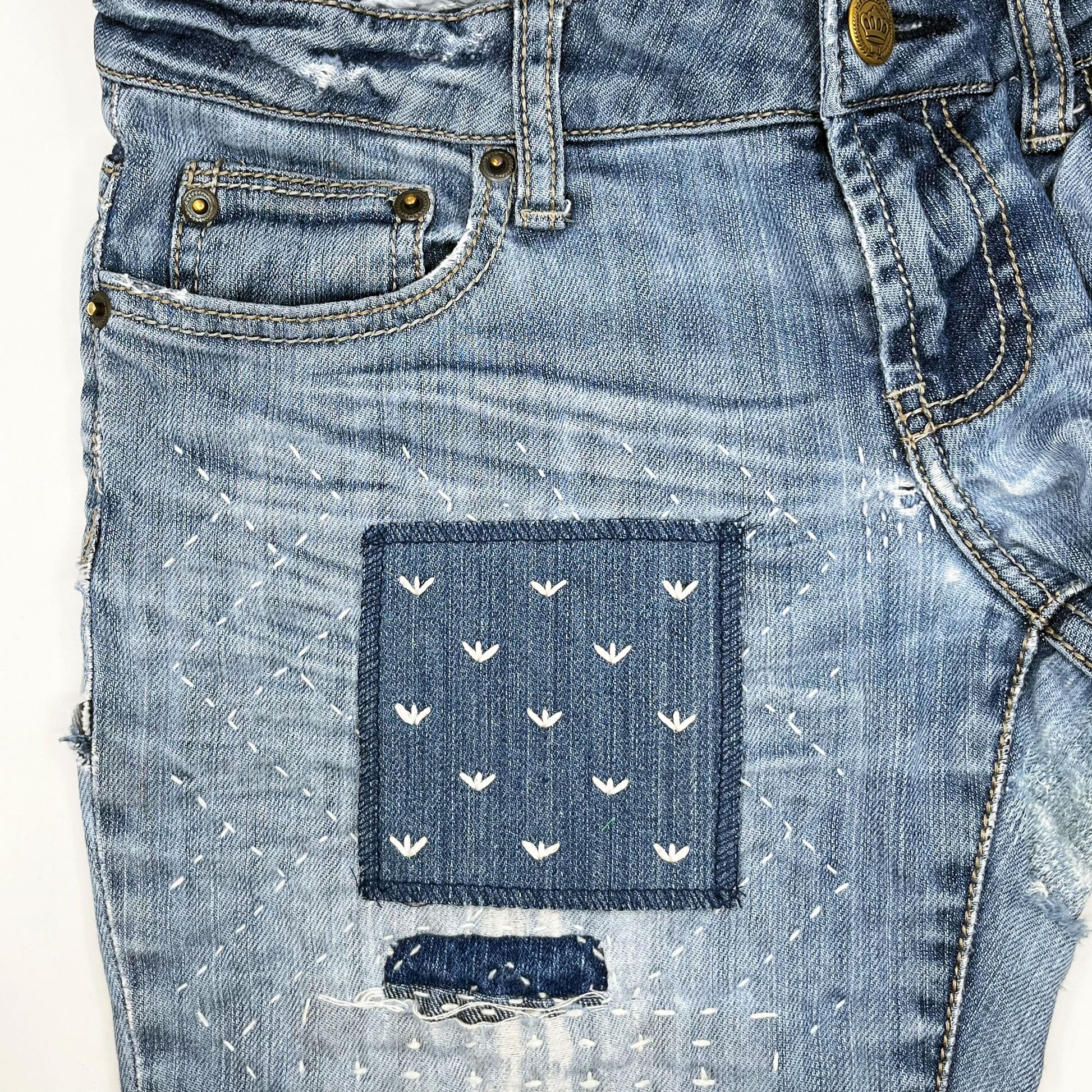 a square denim patch, with ivory stitches that look like birds feet or sprouts spread out in a diamond pattern, on a pair of jeans with other visible mends and sashiko stitching