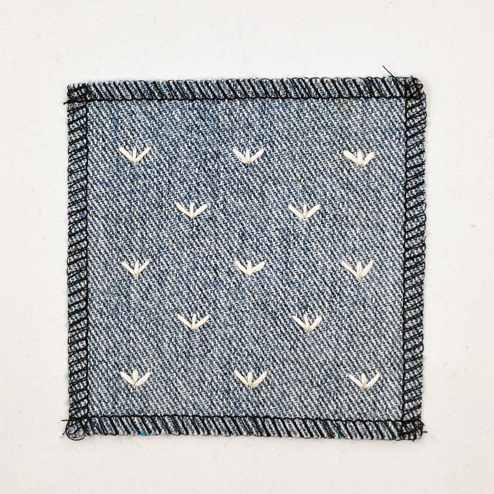 a square denim patch, with ivory stitches that look like birds feet or sprouts spread out in a diamond pattern, with overlocked edges
