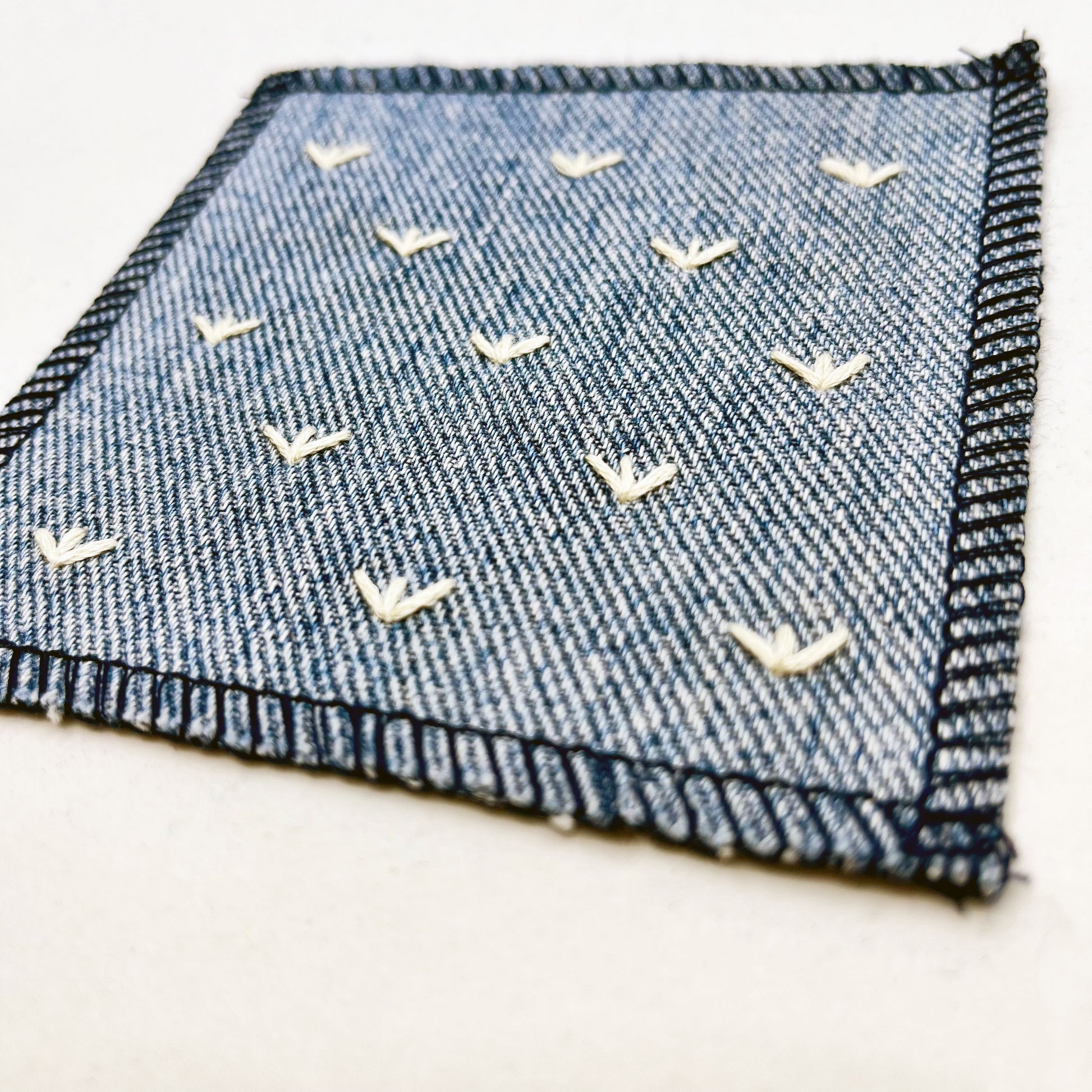 a close up angled view of a square denim patch, with ivory stitches that look like birds feet or sprouts spread out in a diamond pattern, with overlocked edges, on a white background