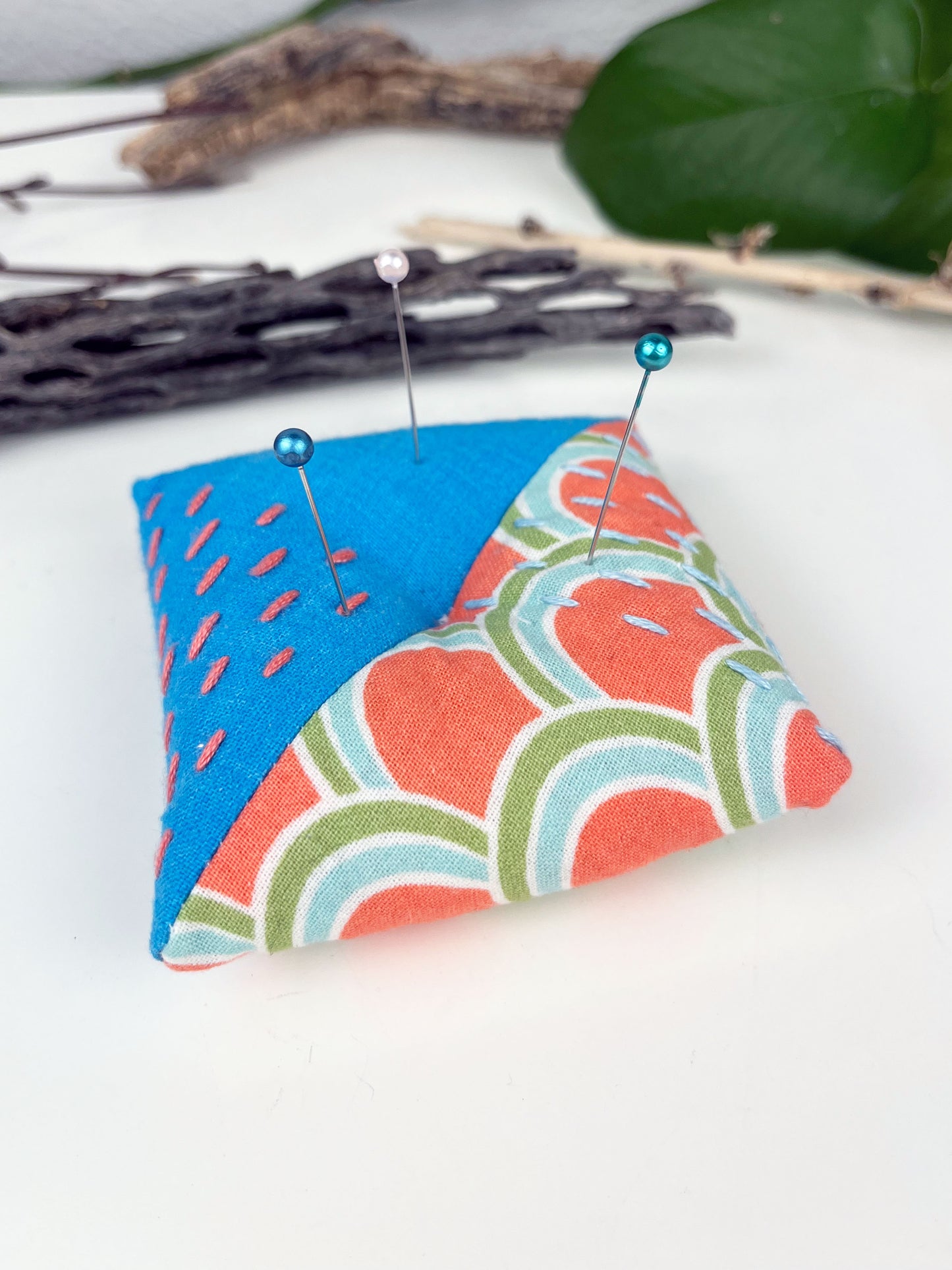 a square pillow shaped pincushion made from bright blue and coral arch patterned fabric, with sashiko stitching in contrasting colors, on a white counter with dried twigs seedpods and a pothos plant in the background
